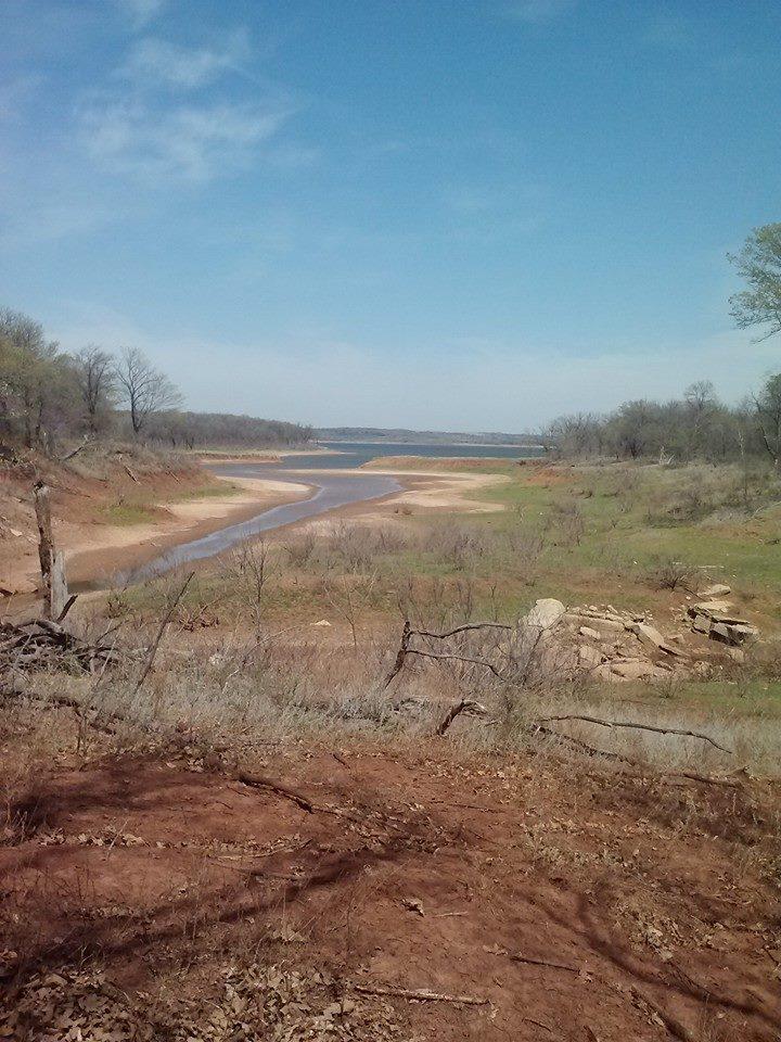 A scenic view of a winding river surrounded by dry land and sparse vegetation under a clear blue sky. The landscape features sandy banks, scattered rocks, and trees in the background, suggesting a tranquil and natural environment. Eagle View, Kaw Lake mountain bike trail.