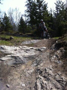 A mountain biker rides down a rocky trail in a forested area, with trees and greenery surrounding the path. The biker is wearing a helmet and is mid-motion, navigating the rugged terrain. Big Sweat (Valley of the Five Lakes / Wabasso) mountain bike trail.