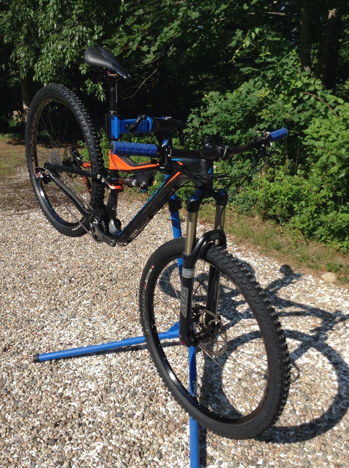Specialized Stumpjumper FSR Expert Carbon EVO 29: A mountain bike on a repair stand, positioned upright with its front wheel raised off the ground, set against a backdrop of greenery. The bike features a black and orange frame with blue accents, and it has thick, knobby tires suitable for off-road use. The stand is blue and made of metal, resting on a gravel surface.