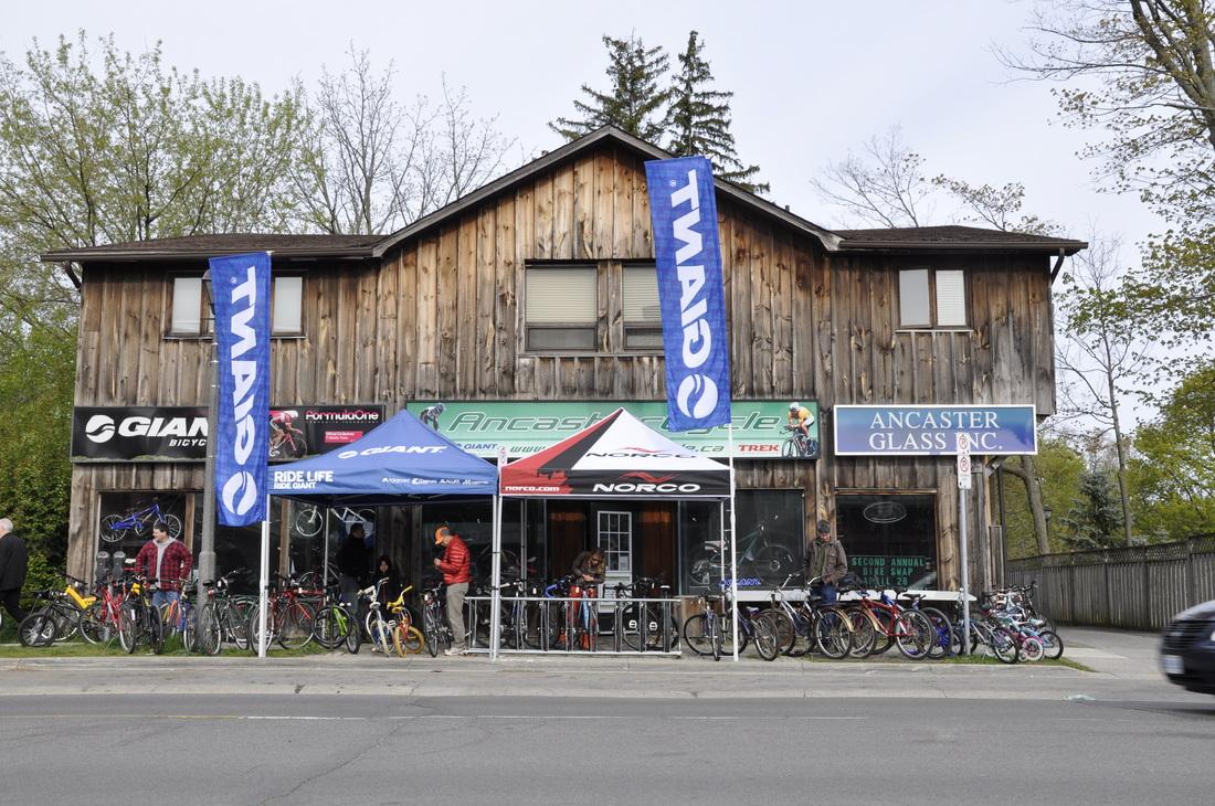 A wooden building featuring multiple bike shop signs, including "Giant" and "Norco," with tents set up in front displaying bicycles. Several people are gathered outside, along with various parked bicycles. Trees and a street are visible in the background.