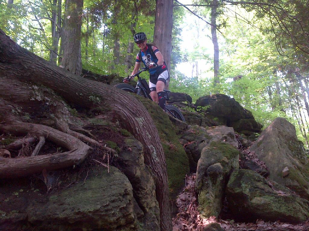 Mountain biker navigating rocky terrain in a wooded area, surrounded by trees. The cyclist is positioned on a steep, moss-covered incline, showcasing an adventurous outdoor activity. Mono Cliffs Provincial Park mountain bike trail.