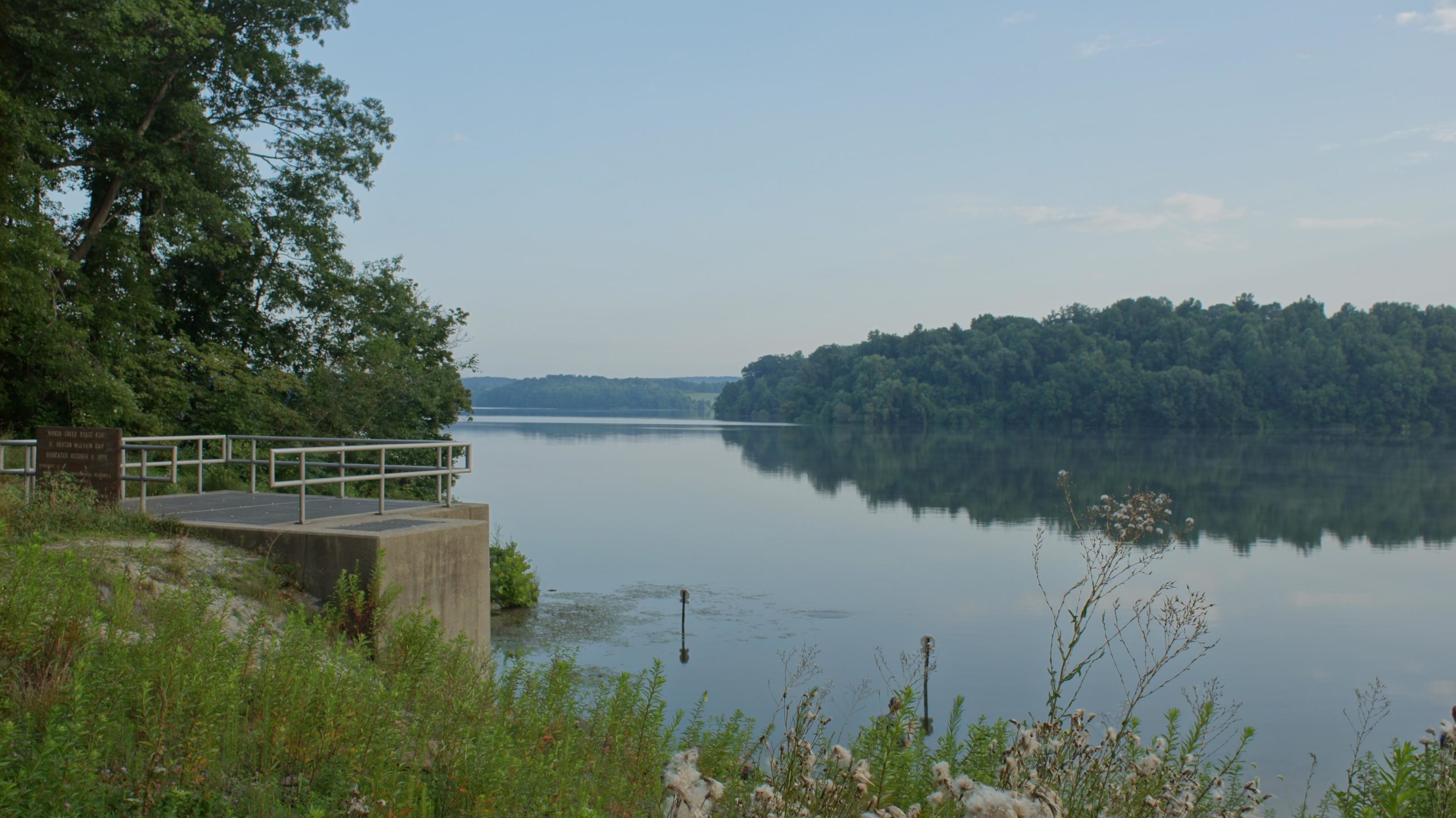 A serene view of a tranquil lake surrounded by lush green trees, featuring a small observation deck with a railing at the water's edge. The calm waters reflect the surrounding landscape, creating a peaceful atmosphere. Soft morning light illuminates the scene, highlighting the natural beauty of the area. Marsh Creek Park mountain bike trail.