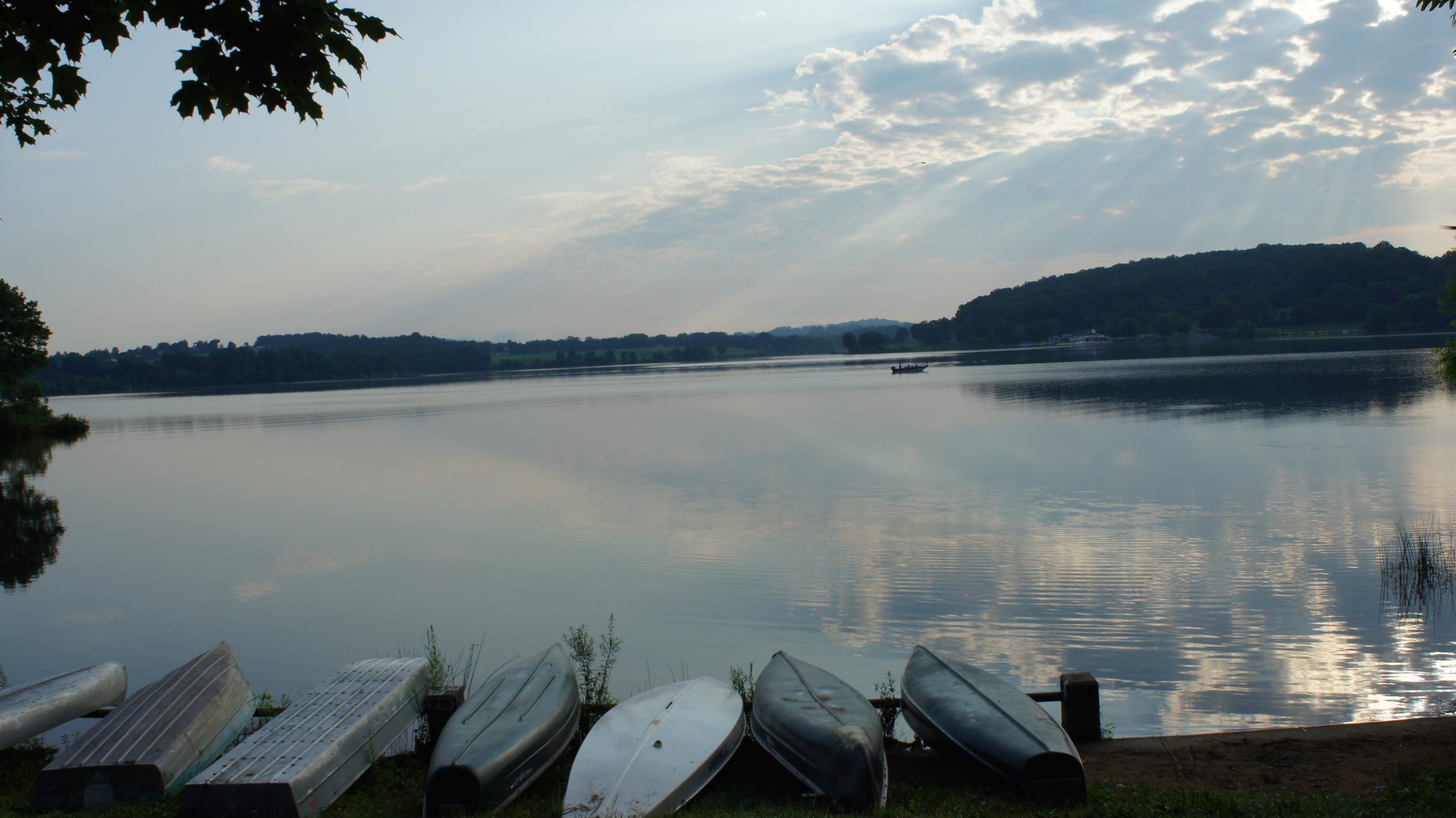 A serene lakeside scene featuring several boats lined up along the shore, with calm water reflecting the cloudy sky. In the distance, a small boat is visible on the water, and rolling green hills surround the lake, creating a peaceful atmosphere. Marsh Creek Park mountain bike trail.
