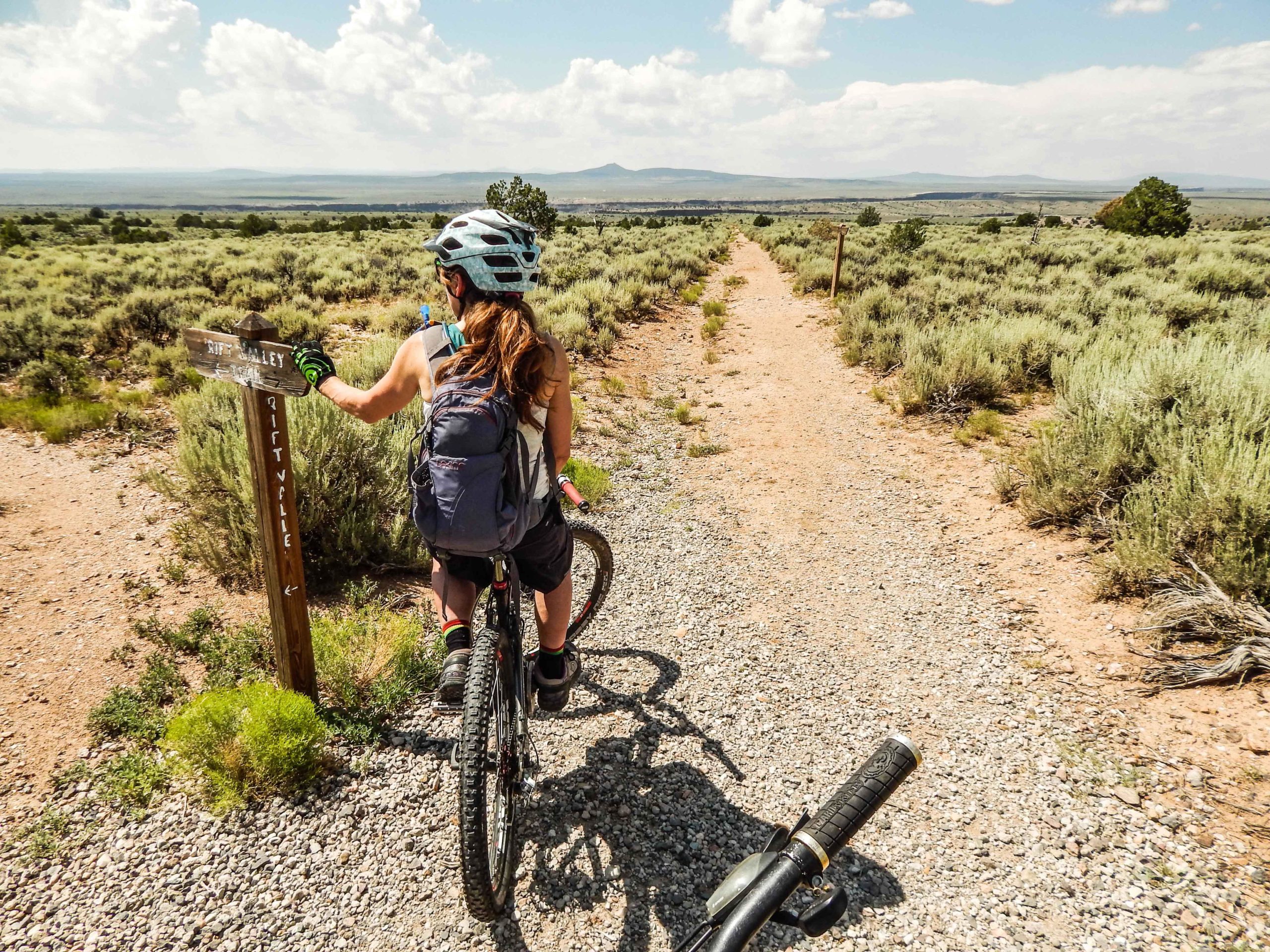 A person on a mountain bike is stopped on a dirt path, looking at a trail sign labeled "Rift Valley." The surrounding landscape features sagebrush and distant hills under a partly cloudy sky. The cyclist is wearing a helmet and a backpack, prepared to continue their ride. Taos Valley Overlook mountain bike trail.