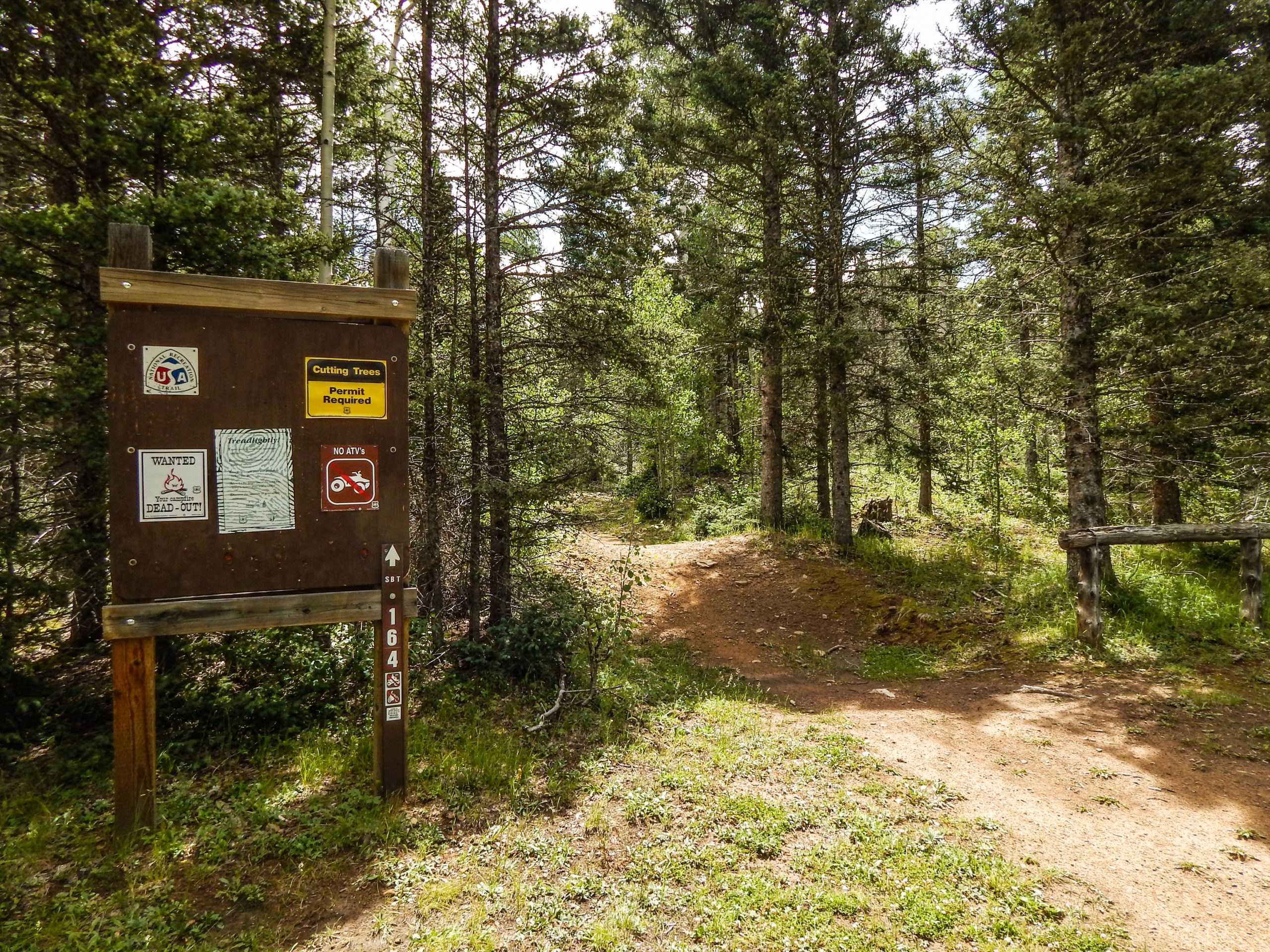 Wooden trail sign in a forested area, indicating rules about tree cutting and permits, with adjacent signs showing no ATVs and a "wanted" notice. A dirt path diverges from the trail, surrounded by greenery and tall trees. South Boundary (164) mountain bike trail.