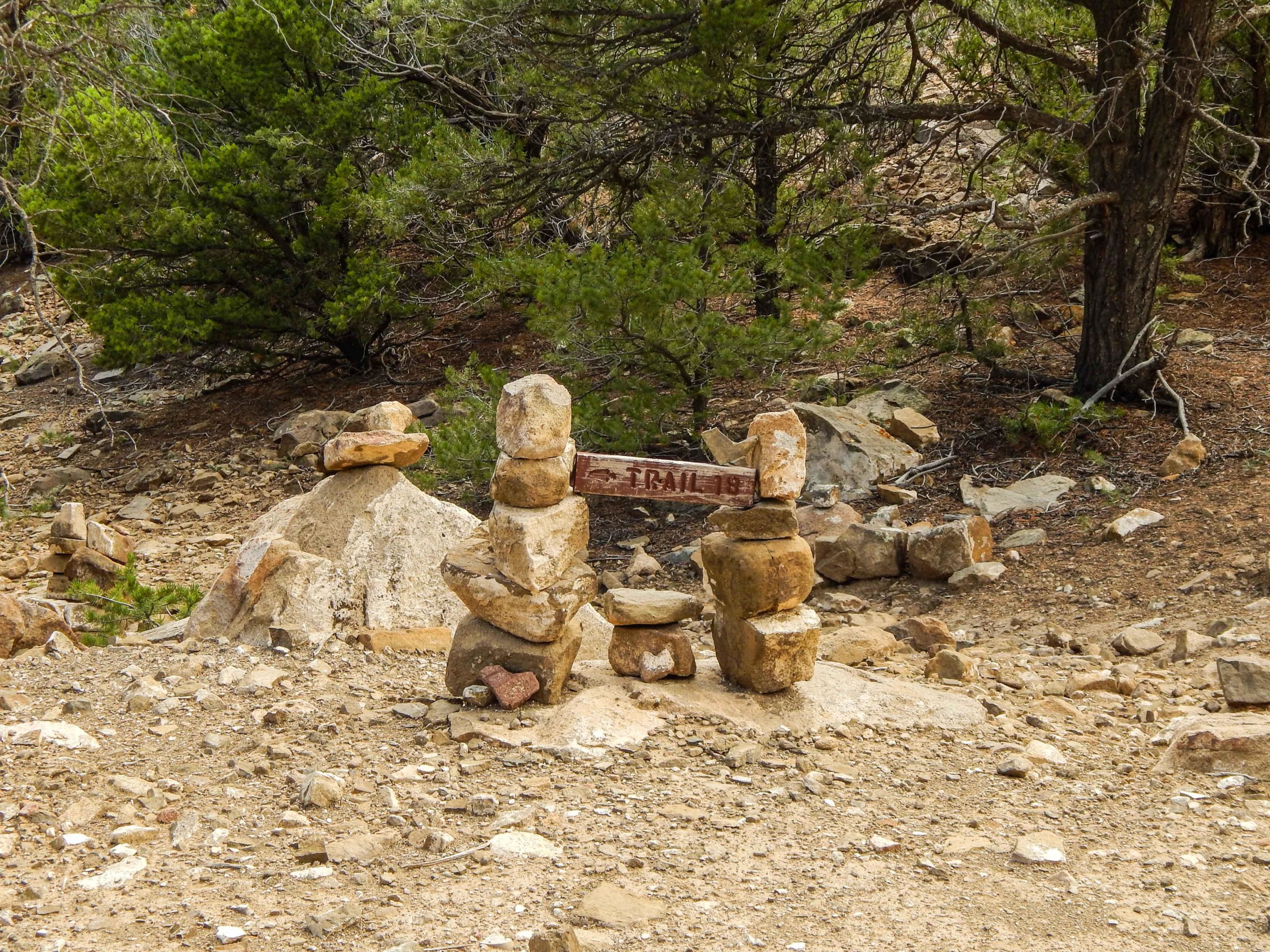 A stacked stone formation serves as a trail marker in a natural setting, surrounded by trees and rocky terrain. The wooden sign, partially obscured by the stones, indicates the trail name. The ground is covered with gravel and small rocks, typical of a hiking path. Trail 18 ("rio Grande Del Rancho") mountain bike trail.
