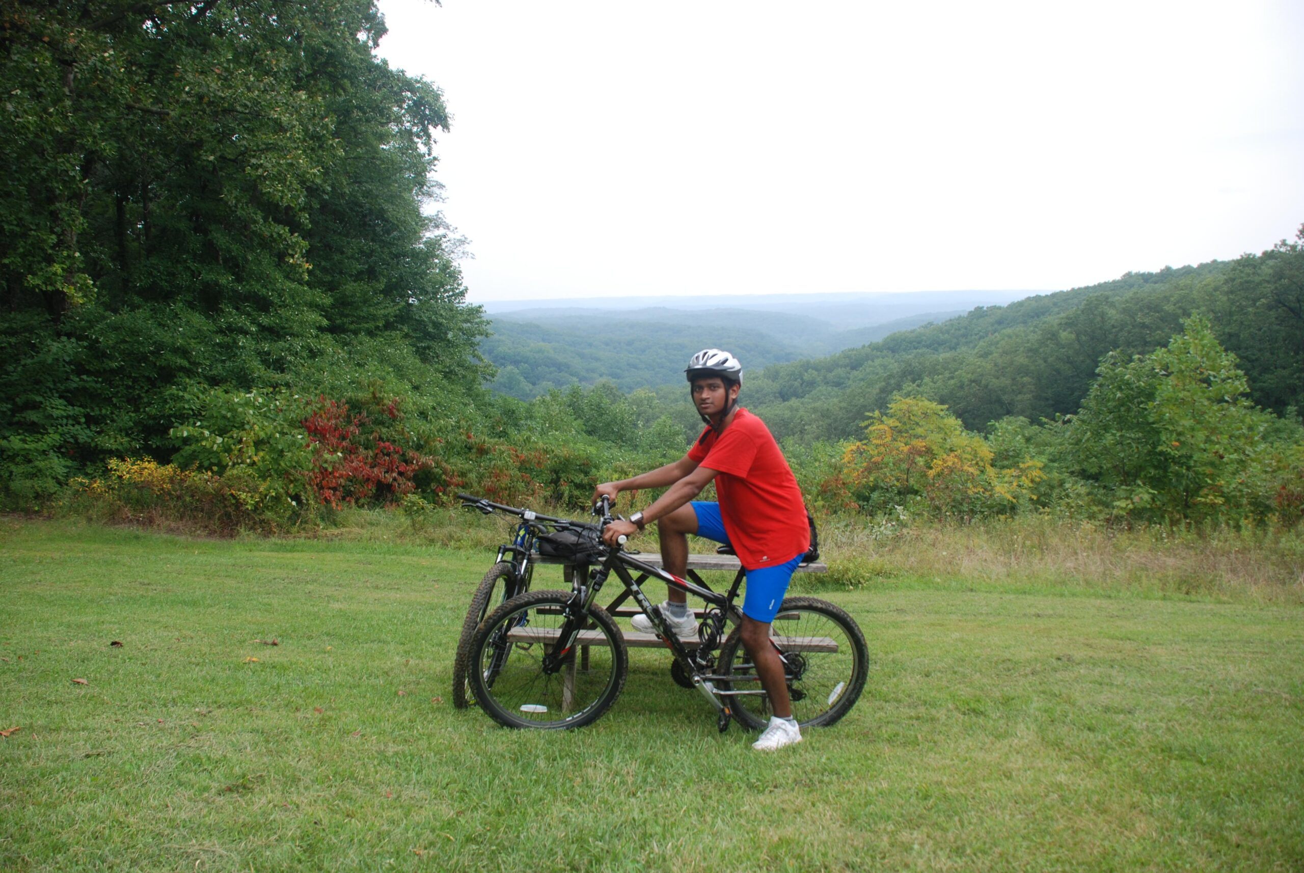 A person wearing a helmet and casual athletic clothing is sitting on a mountain bike, with one foot on the ground and the other on the bike's frame. They are positioned in a grassy area overlooking a scenic landscape of rolling hills and trees under a cloudy sky. A second bike is propped nearby, and there are patches of colorful foliage in the background. Brown County Park mountain bike trail.