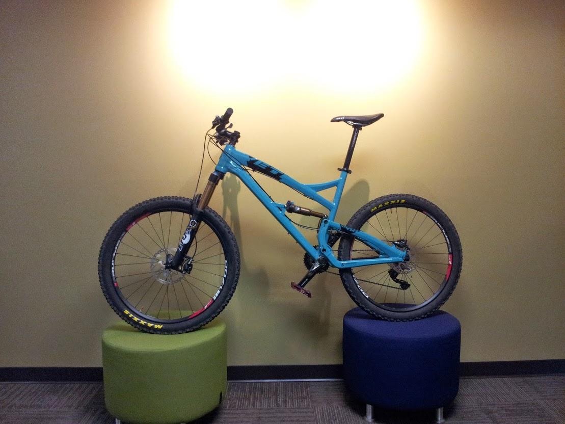 Yeti SB66: A blue mountain bike displayed on two cylindrical stools against a plain green wall, with a light illuminating the bike from above. The bike features thick tires and a modern design, emphasizing its suitability for off-road riding.