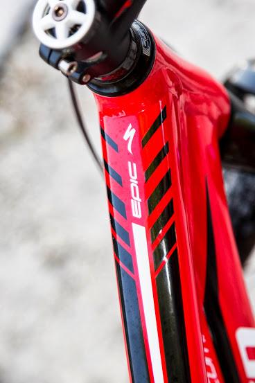 Specialized Epic Comp 29er: Close-up image of a red bicycle frame featuring sleek black stripes and the word "Epic" prominently displayed. The frame is angled to show the handlebar and the upper portion of the bike, highlighting its sporty design and attention to detail.