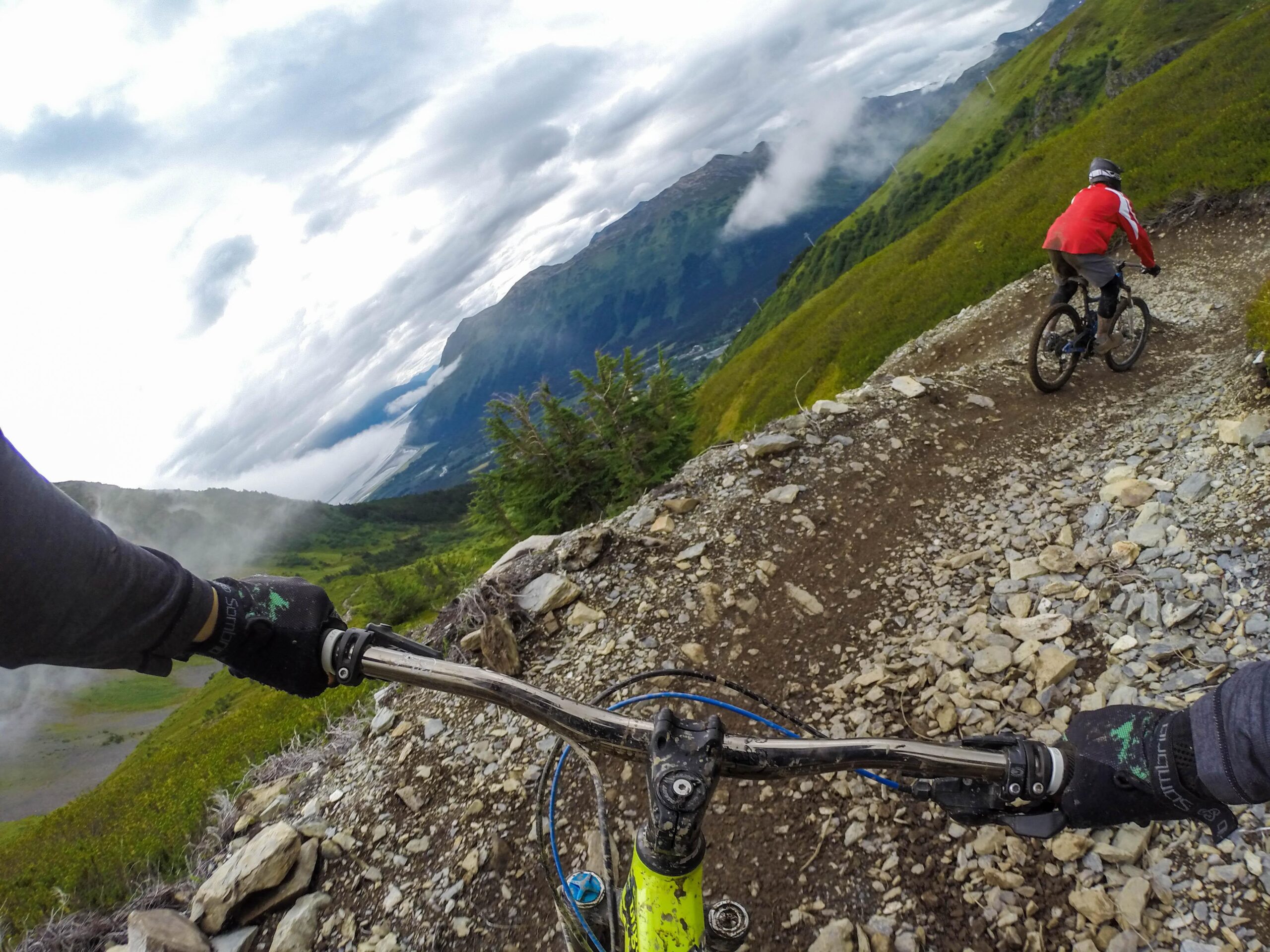 Mountain bikers navigating a rocky trail on a hillside, with a panoramic view of green landscapes and clouds in the background. The image captures the perspective of the rider in the foreground and another cyclist visible in the distance. Alyeska Downhill mountain bike trail.