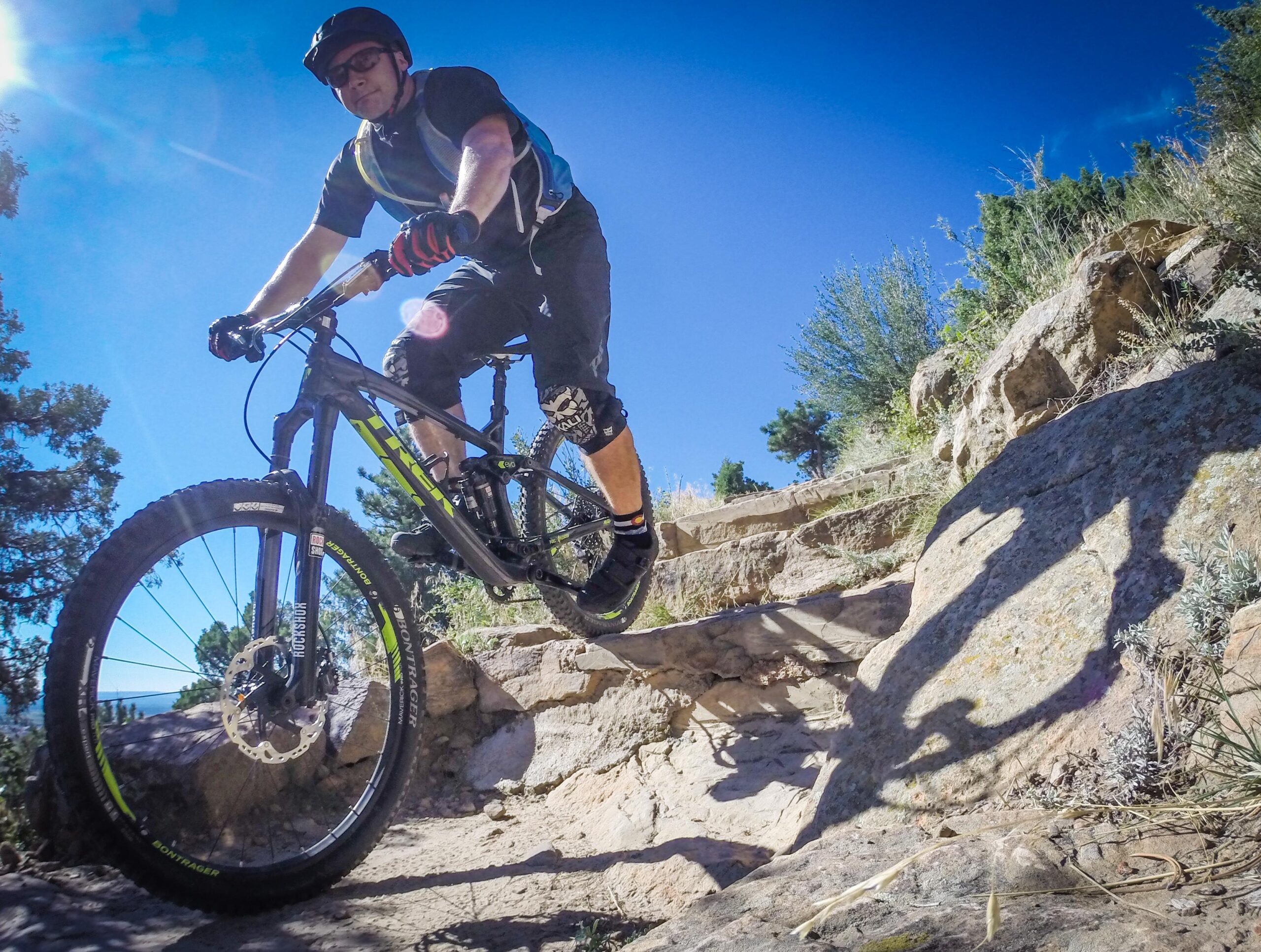 A mountain biker navigating a rocky trail under a clear blue sky. The rider wears a helmet and protective gear, showcasing an athletic posture as they maneuver over the uneven terrain. Sunlight creates a bright glare, highlighting the dynamic action of the ride. Red Rocks / Dakota Ridge mountain bike trail.