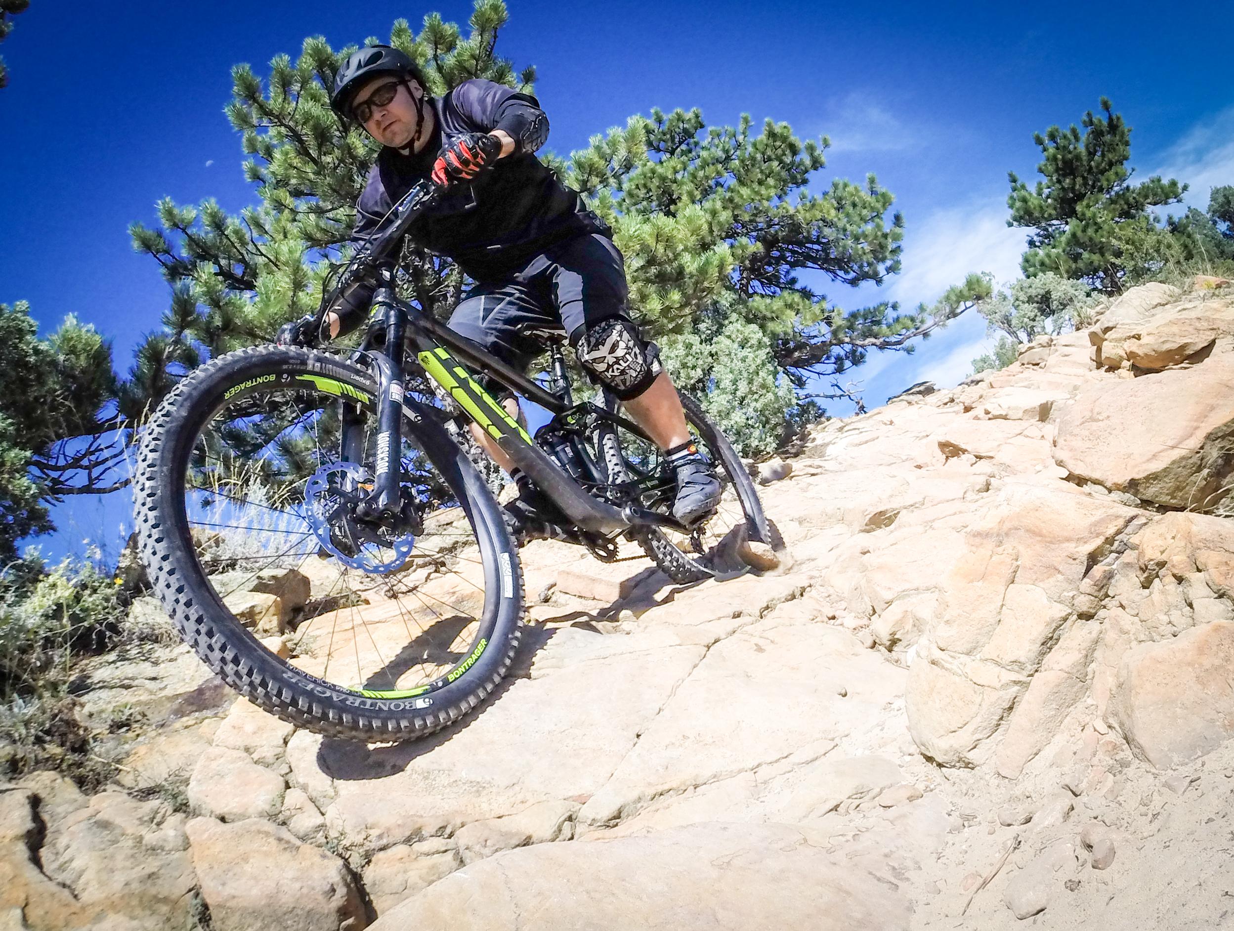 A mountain biker navigating a rocky trail under a blue sky, surrounded by pine trees. The cyclist is wearing a helmet and protective gear while leaning into the terrain. Red Rocks / Dakota Ridge mountain bike trail.
