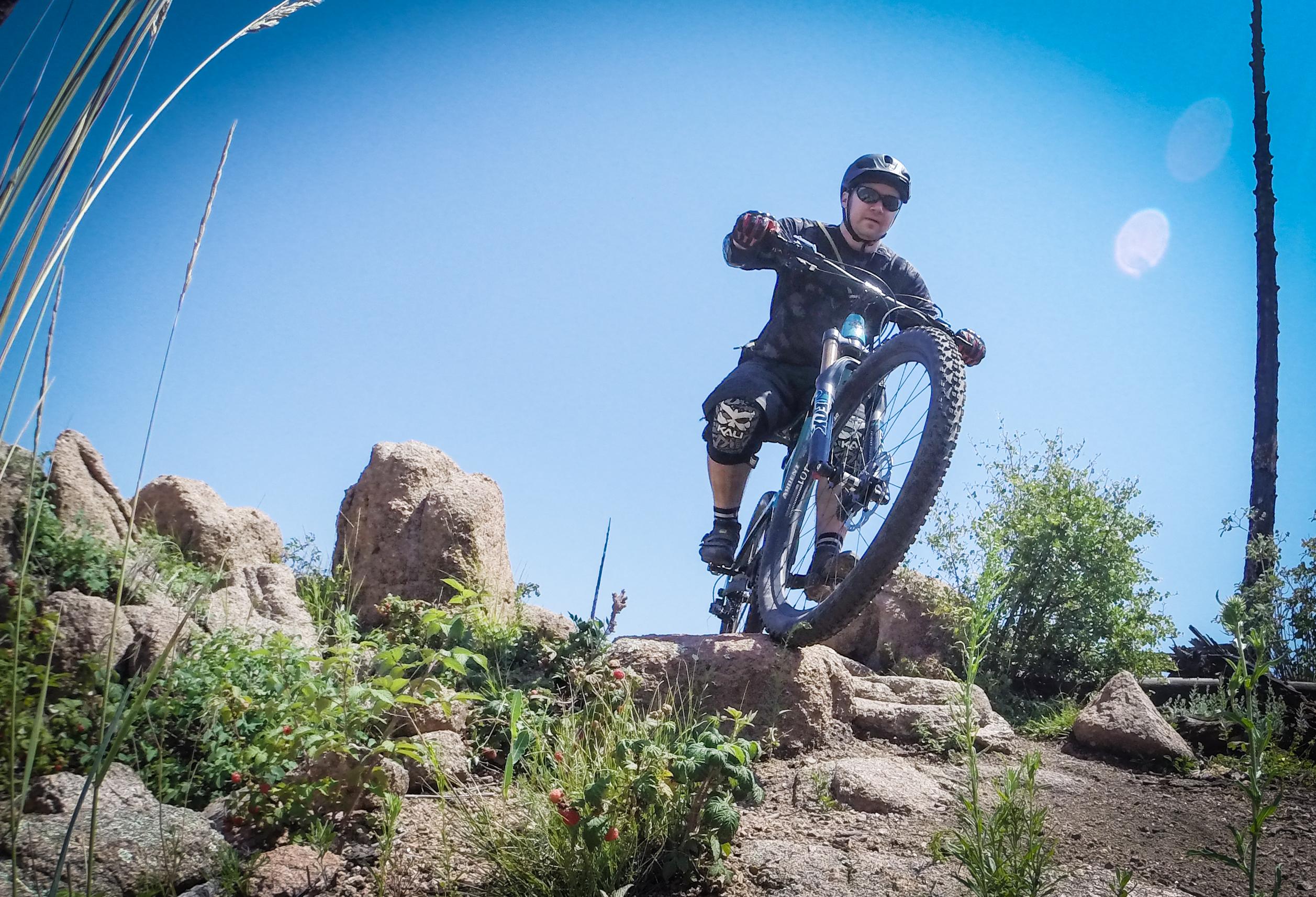A mountain biker navigating a rocky trail surrounded by greenery, set against a clear blue sky. The rider is wearing a helmet and protective gear, riding downhill on a mountain bike. Buffalo Creek mountain bike trail.