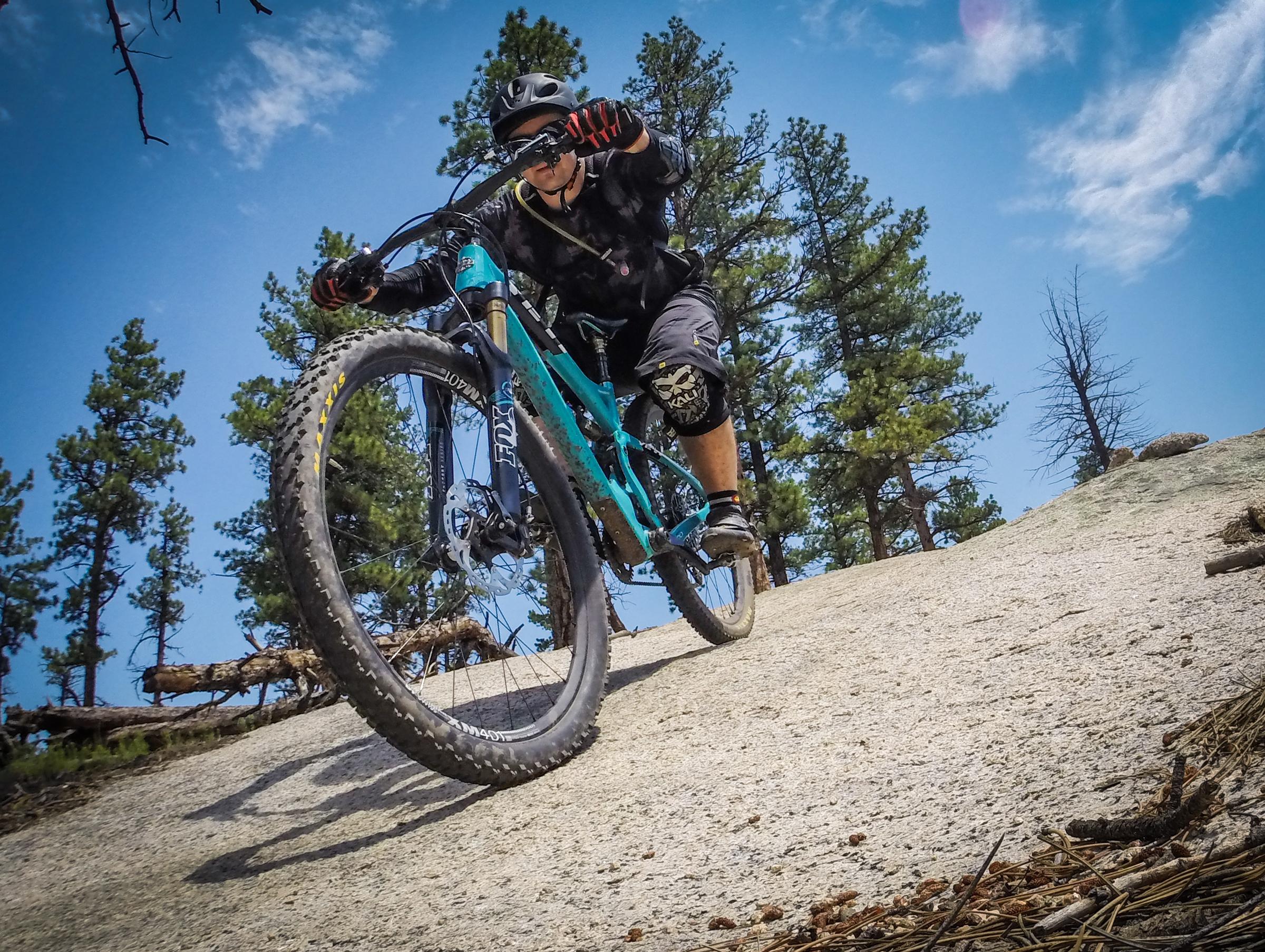 A mountain biker riding downhill on a rocky trail, surrounded by tall pine trees under a blue sky. The rider is wearing a helmet and protective gear, demonstrating skill and control as they navigate the terrain. Buffalo Creek mountain bike trail.