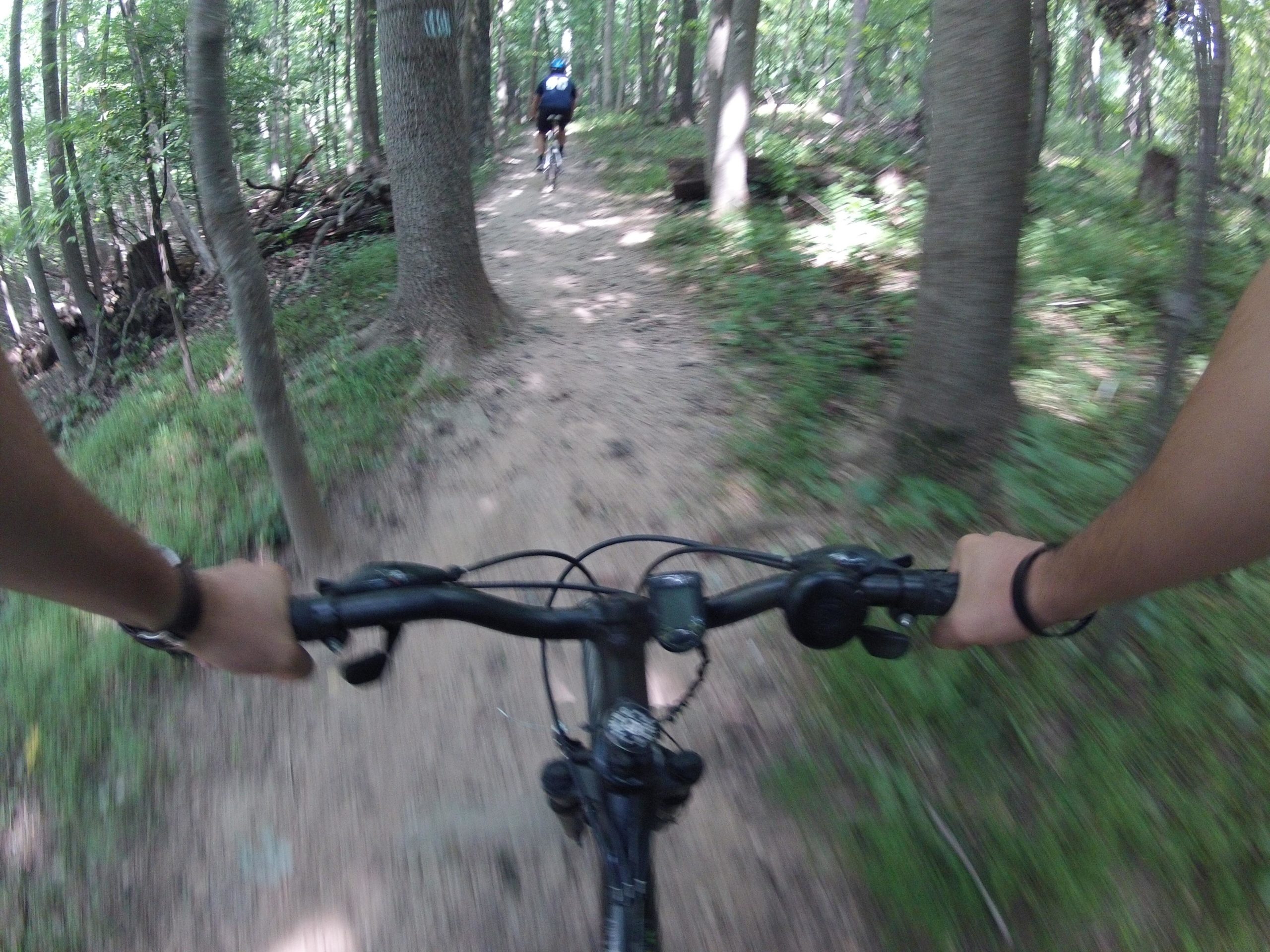 A cyclist's perspective while riding on a muddy trail through a lush green forest. The foreground shows hands gripping the bike handlebars, with trees and fellow cyclists blurred in the background, suggesting motion and an adventurous outdoor activity. Cabin John Trail mountain bike trail.
