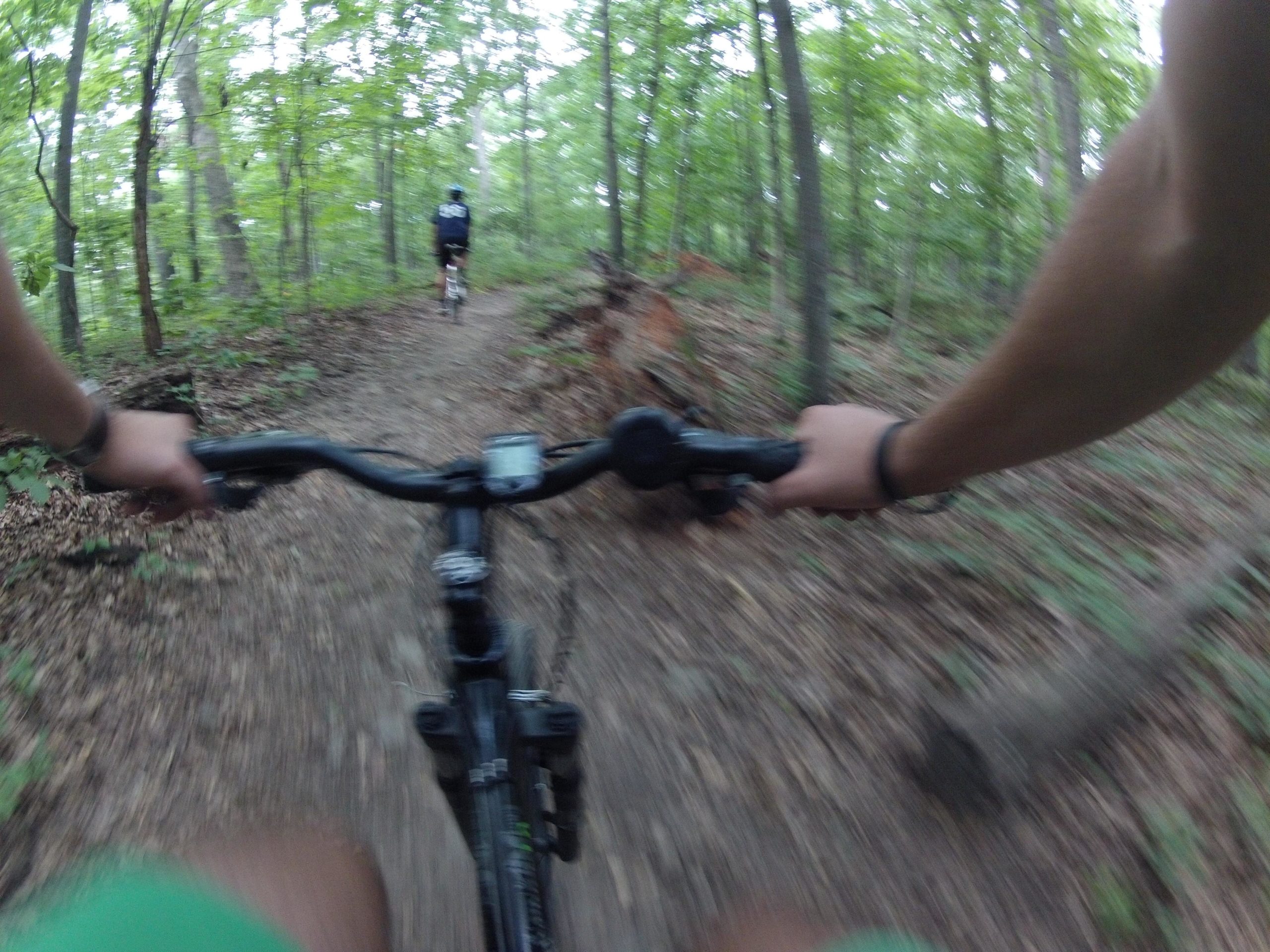 A cyclist's view of a dirt trail in a wooded area, with trees and greenery on either side. The handlebars of the bike are in the foreground, and a second cyclist is visible in the background, riding down the path. The image conveys a sense of movement and adventure in nature. Cabin John Trail mountain bike trail.