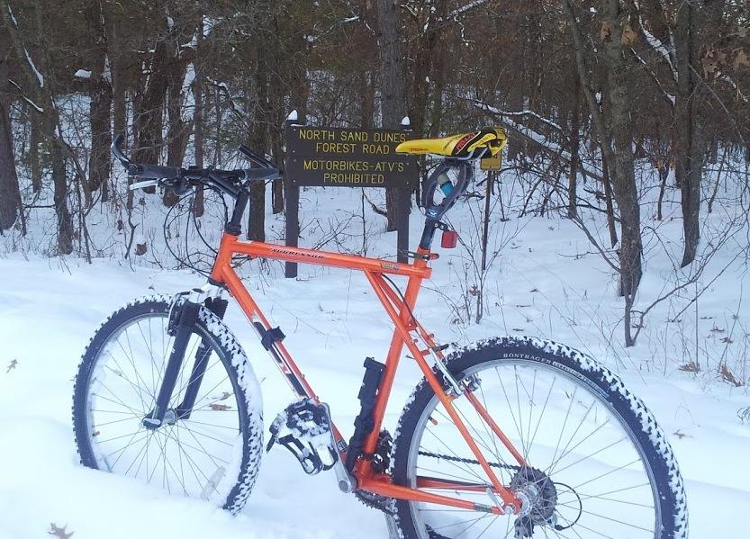 GT Aggressor 1998, Orange & Black: An orange mountain bike stands in a snowy forest setting, next to a sign that reads "NORTH SAND DUNES FOREST ROAD - MOTORBIKES - ATVs PROHIBITED." The bike is partially covered with snow, and the surrounding trees are bare, indicating winter conditions.