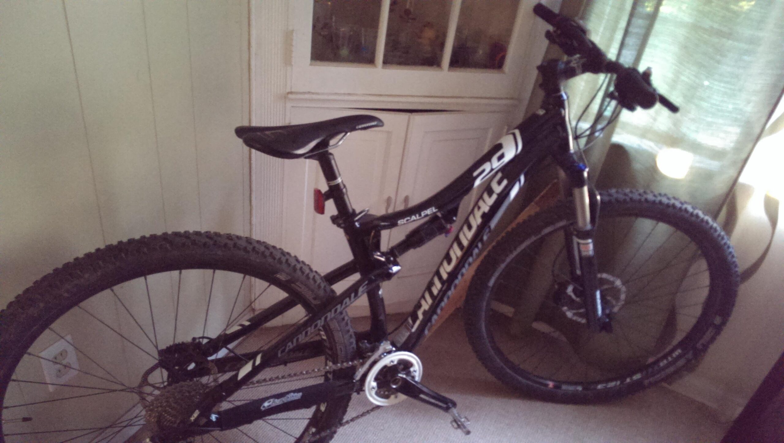 A black Cannondale Scalpel mountain bike leaning against a wall indoors, with a partially visible door and green curtains in the background. The bike features large, knobby tires and a visible gear system.