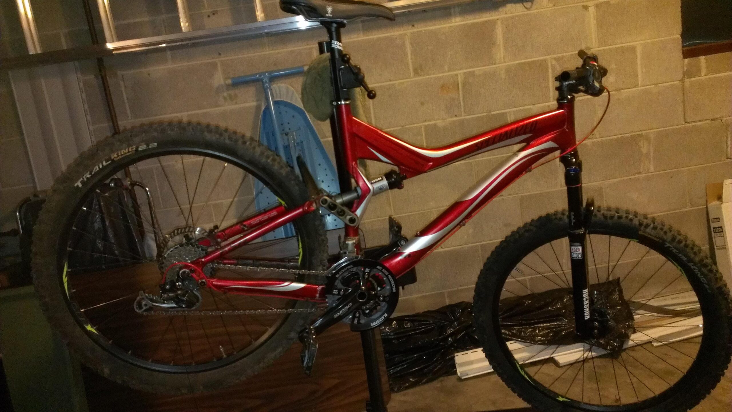 Specialized Stumpjumper FSR Elite: A red mountain bike with a sleek frame and thick tires is mounted on a repair stand in a dimly lit garage. The bike features a dual suspension system and a visible rear derailleur. Various tools and equipment are partially visible in the background, along with a concrete wall.