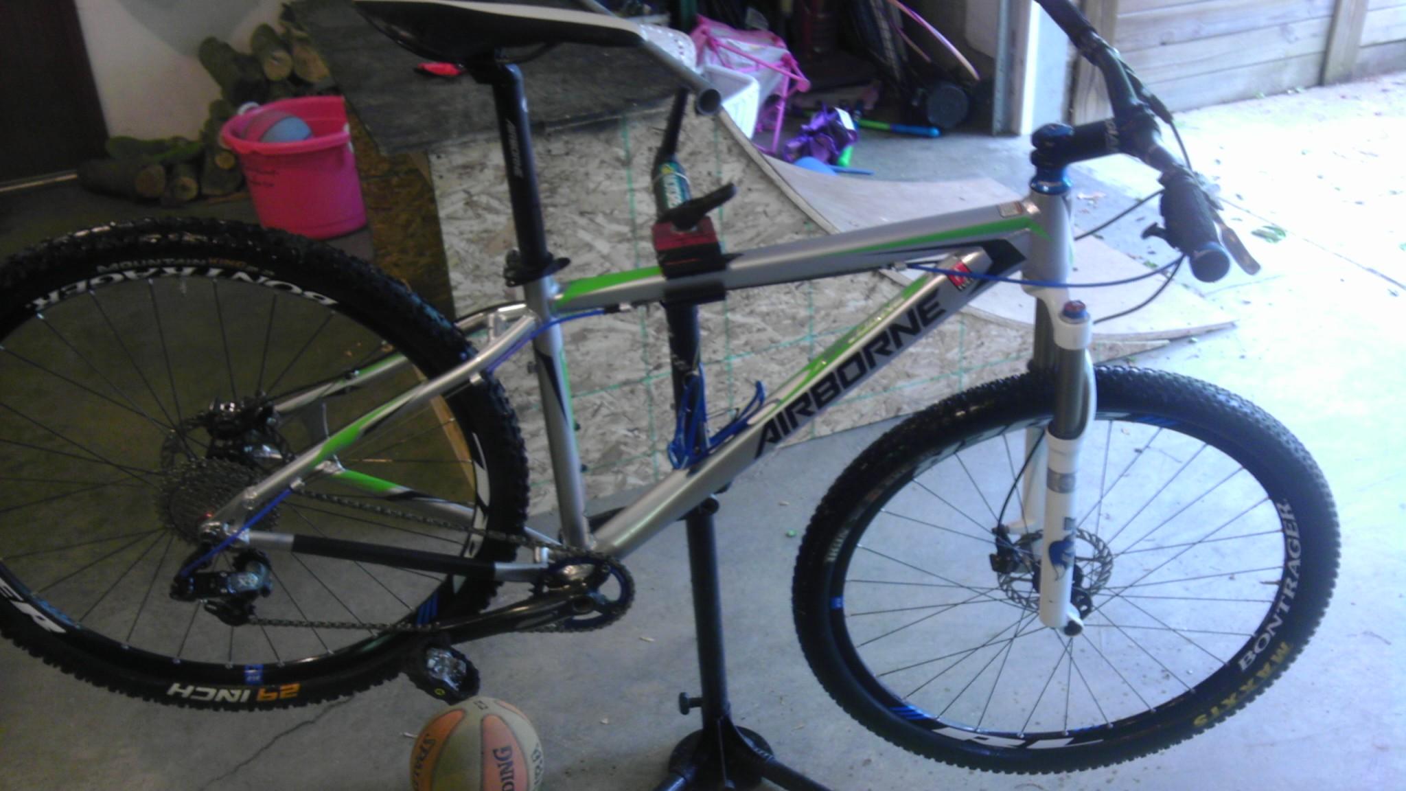 Airborne Goblin: A mountain bike with a silver frame labeled "Airborne" is mounted on a repair stand. The bike features thick tires designed for off-road terrain, and a visible front suspension fork. In the background, various objects, including a pink basket and a ball, can be seen on the garage floor.