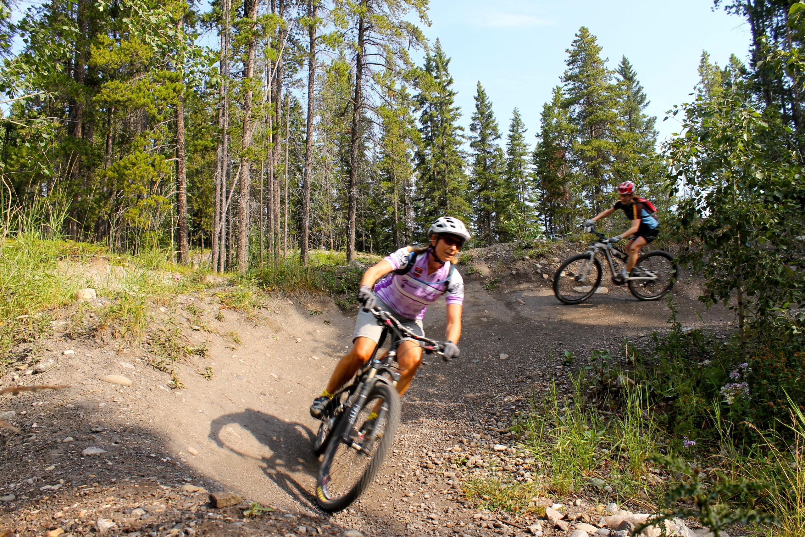 Two mountain bikers riding on a dirt trail through a forested area. The first biker, wearing a purple jersey and helmet, is leaning into a turn with a smile. The second biker, in a red helmet and darker clothing, is slightly behind, also navigating the trail. Tall trees and greenery surround the scene under a clear sky. Canmore Nordic Centre mountain bike trail.