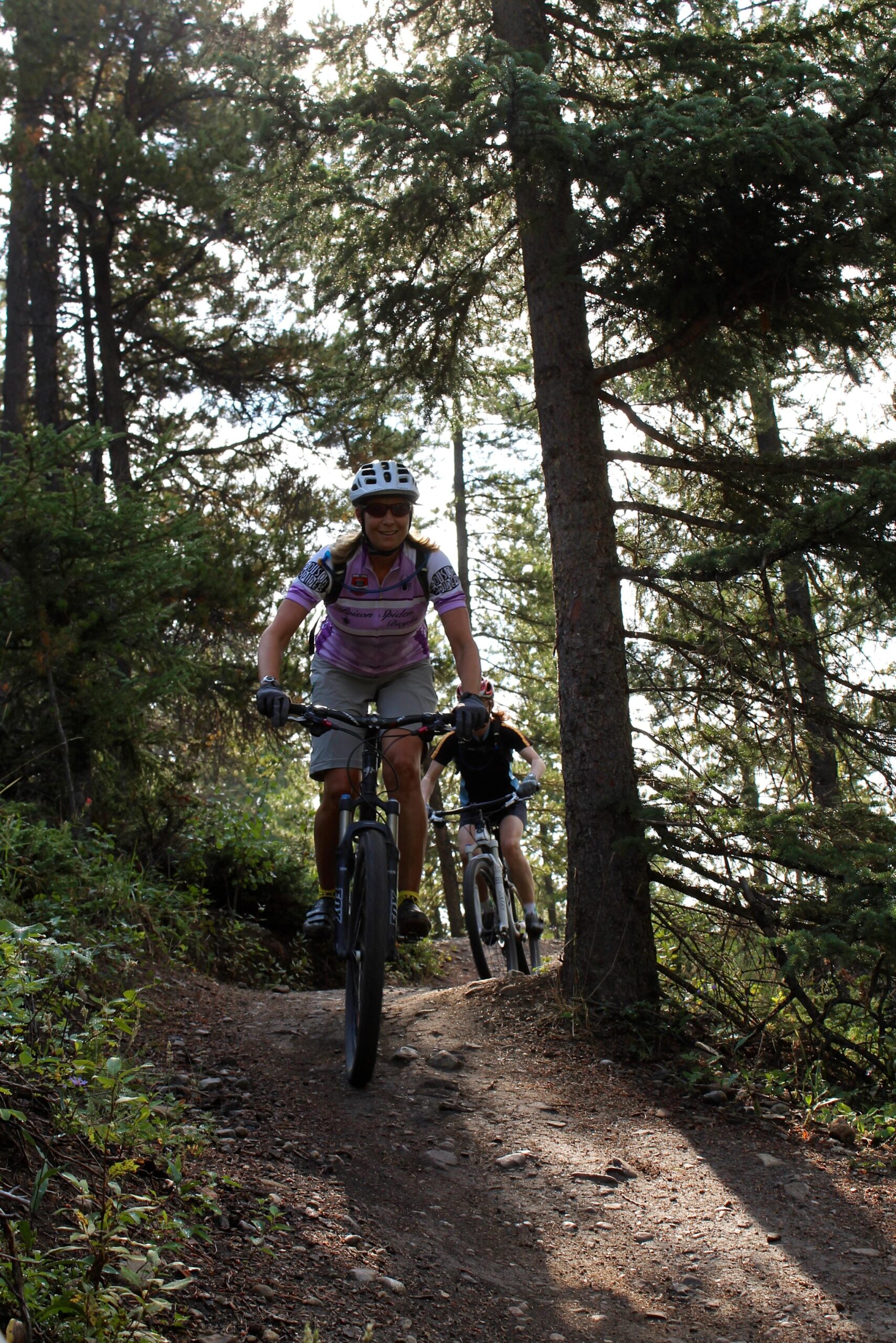 Two mountain bikers riding along a narrow dirt trail surrounded by tall trees and greenery. The first rider, wearing a purple shirt and helmet, smiles as they navigate the trail, while the second rider follows closely behind. The scene is illuminated by natural light filtering through the tree branches. Canmore Nordic Centre mountain bike trail.