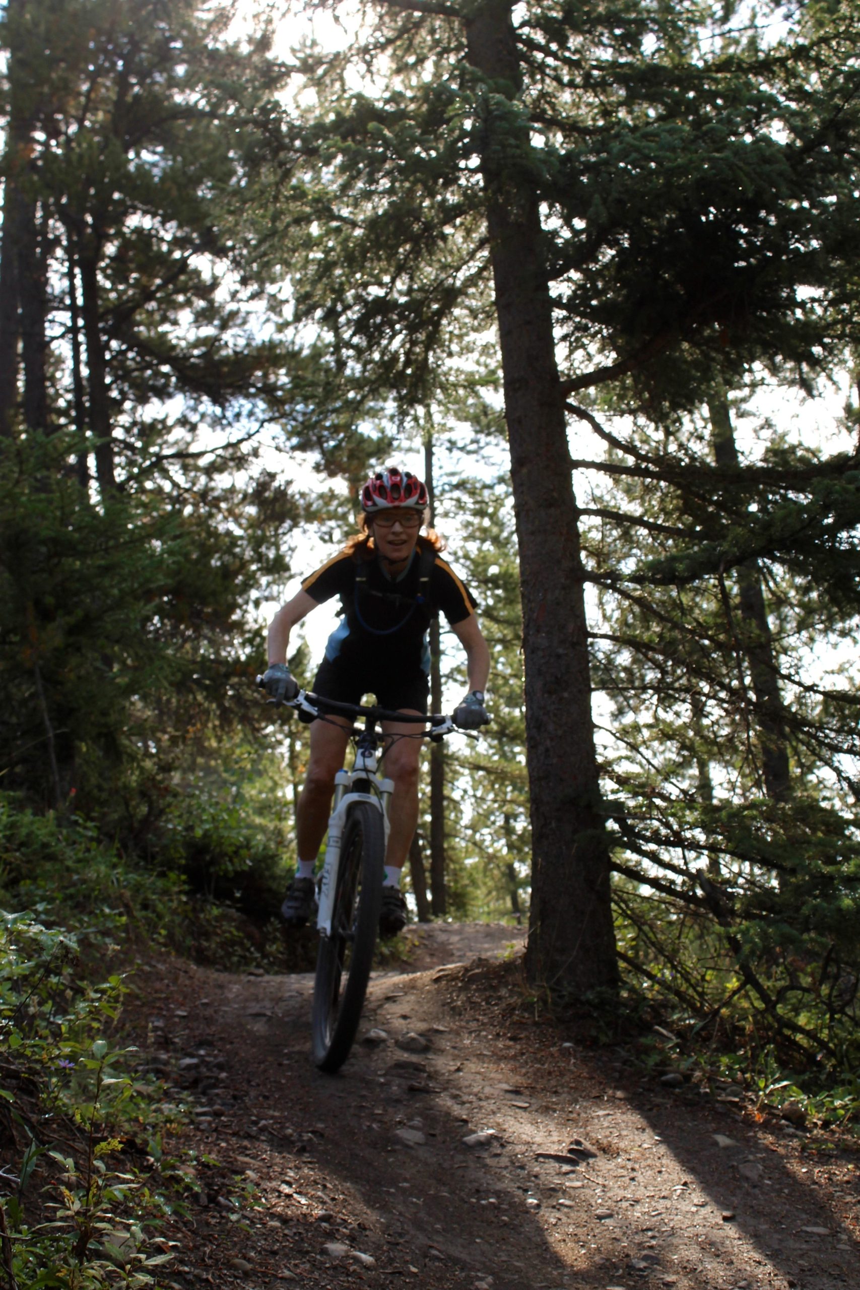 A mountain biker is riding on a dirt trail through a wooded area, surrounded by tall trees and sunlight filtering through the leaves. The rider is wearing a red helmet and glasses, with a focused expression, as they navigate the path. Canmore Nordic Centre mountain bike trail.