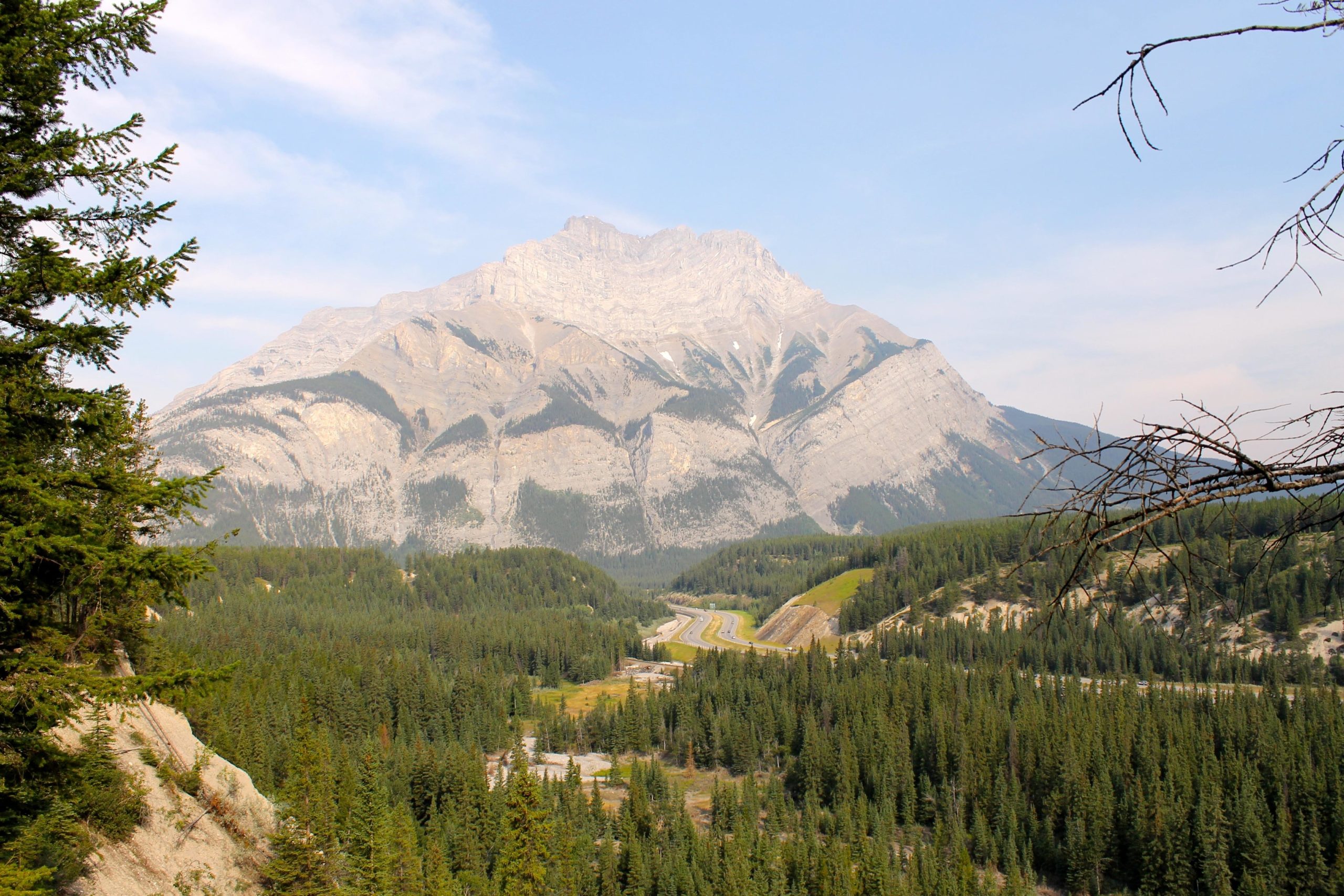 A mountainous landscape featuring a prominent peak in the background, surrounded by dense green forests and a winding road visible through the valley. The sky is partially cloudy, adding depth to the serene natural setting. Tunnel Mountain Trail System mountain bike trail.