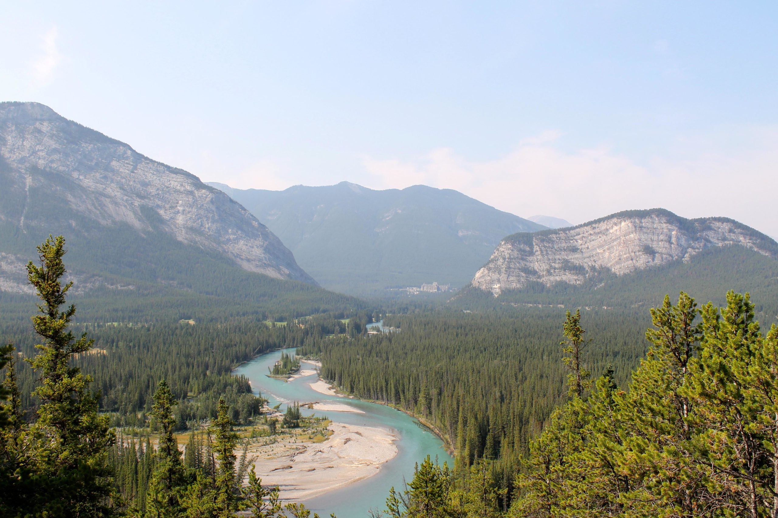 A panoramic view of a winding river flowing through a lush green forest, surrounded by towering mountains under a clear blue sky. The landscape features a mix of evergreen trees, rocky terrain, and visible sandy banks along the river. Tunnel Mountain Trail System mountain bike trail.