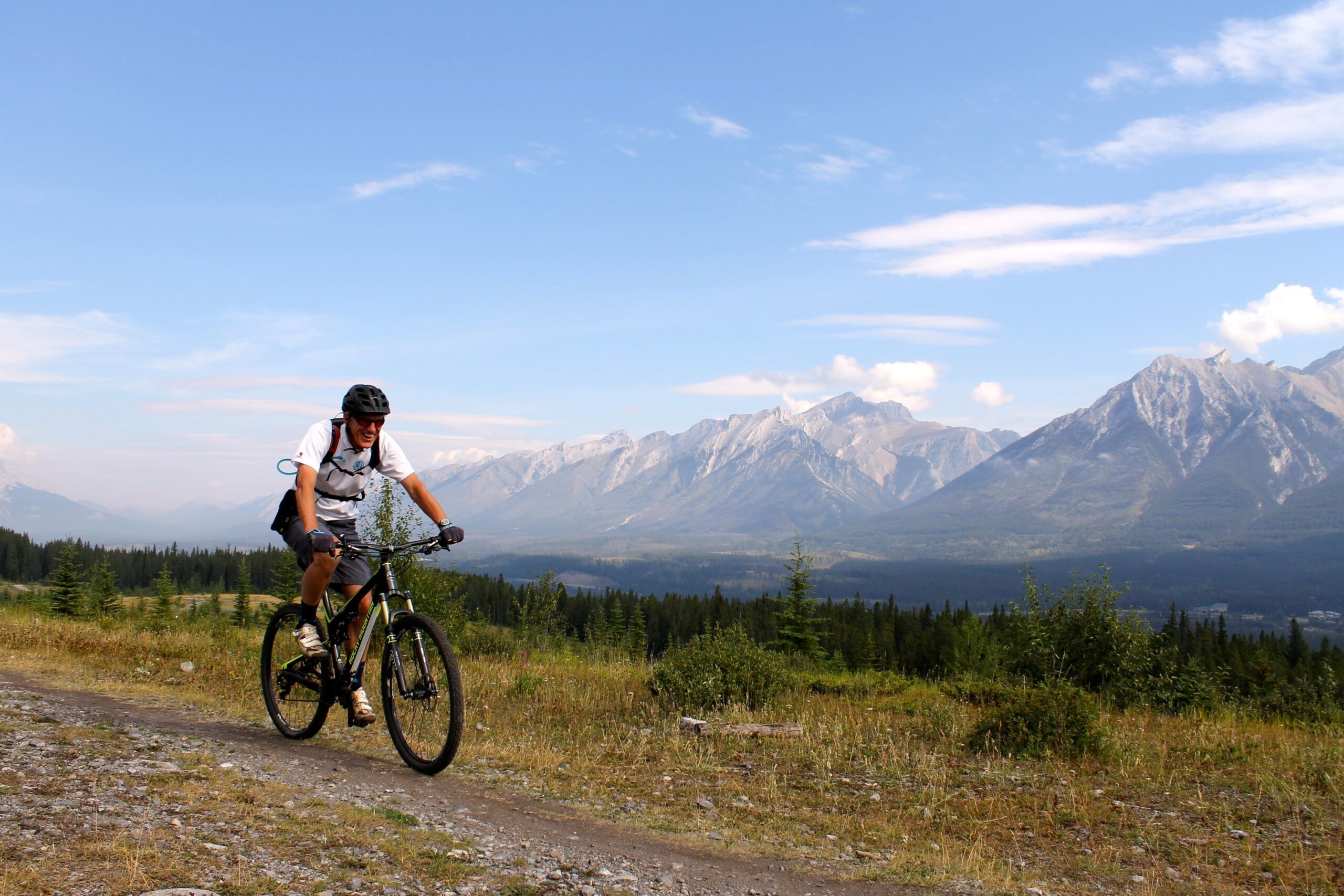 A mountain biker riding along a gravel trail in a mountainous landscape, with trees and fields in the foreground and a clear blue sky above. Majestic mountains can be seen in the background, enhancing the scenic beauty of the outdoor environment. Canmore Nordic Centre mountain bike trail.