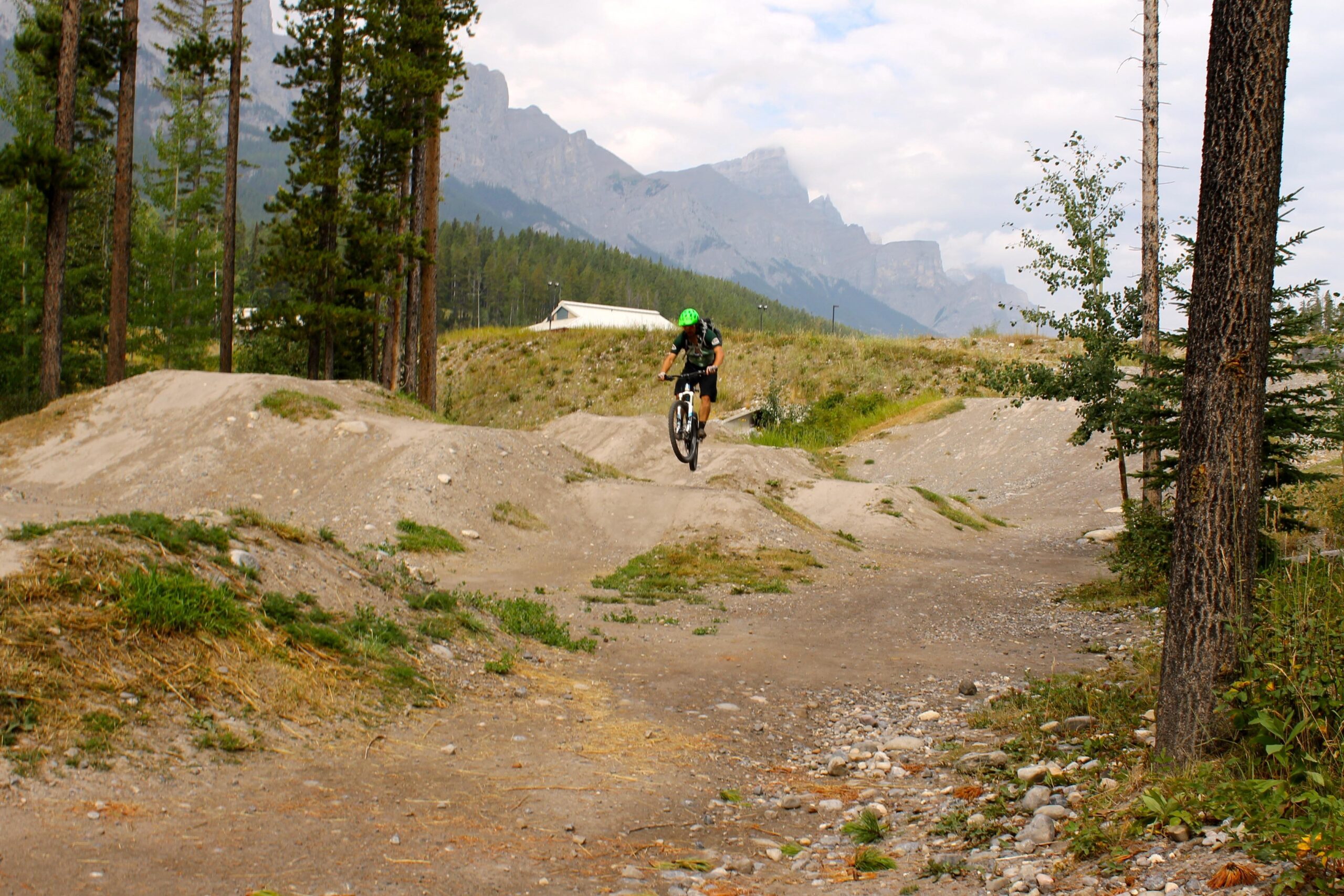 A mountain biker in a green helmet performs a jump on a dirt trail surrounded by trees, with mountains and a cloudy sky in the background. Canmore Nordic Centre mountain bike trail.