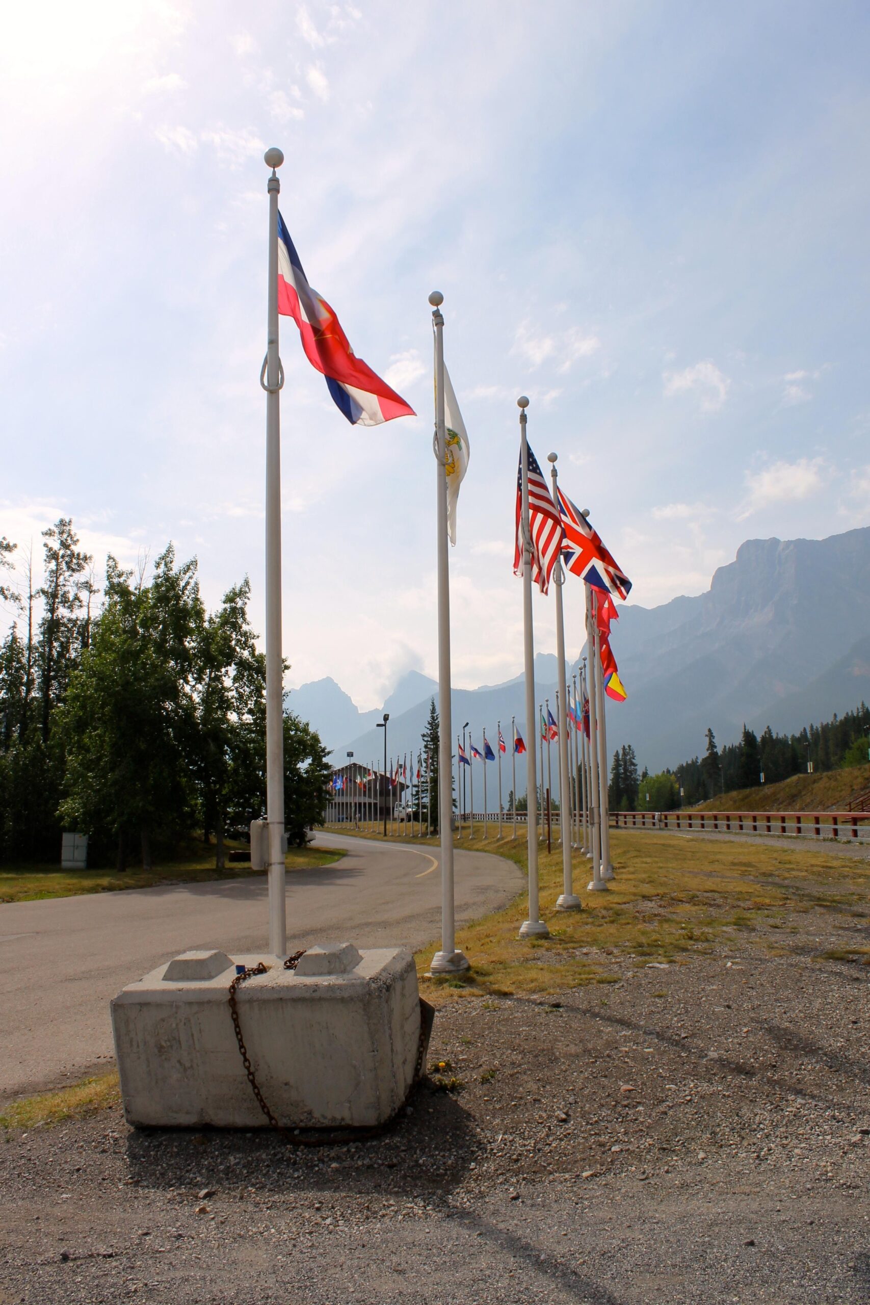 A line of flags from various countries stands along a roadside, with trees and mountains in the background under a partly cloudy sky. In the foreground, a large concrete block is secured with a chain, and a curve in the road leads off into the distance. Canmore Nordic Centre mountain bike trail.