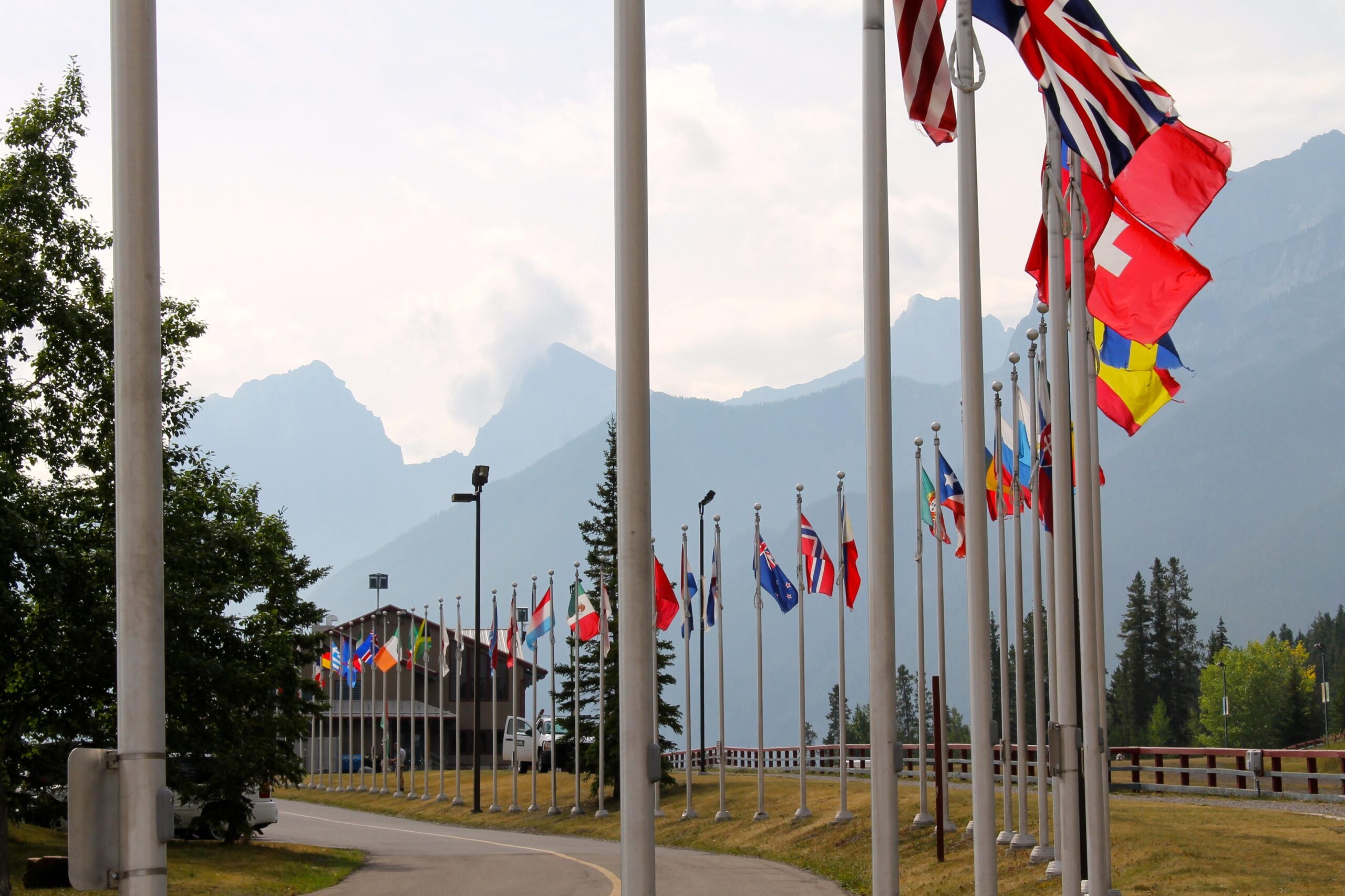 A row of international flags displayed along a pathway, with a backdrop of mountains under a hazy sky. In the background, a building is partially visible, surrounded by greenery and trees. The scene conveys a sense of unity and diversity among nations. Canmore Nordic Centre mountain bike trail.