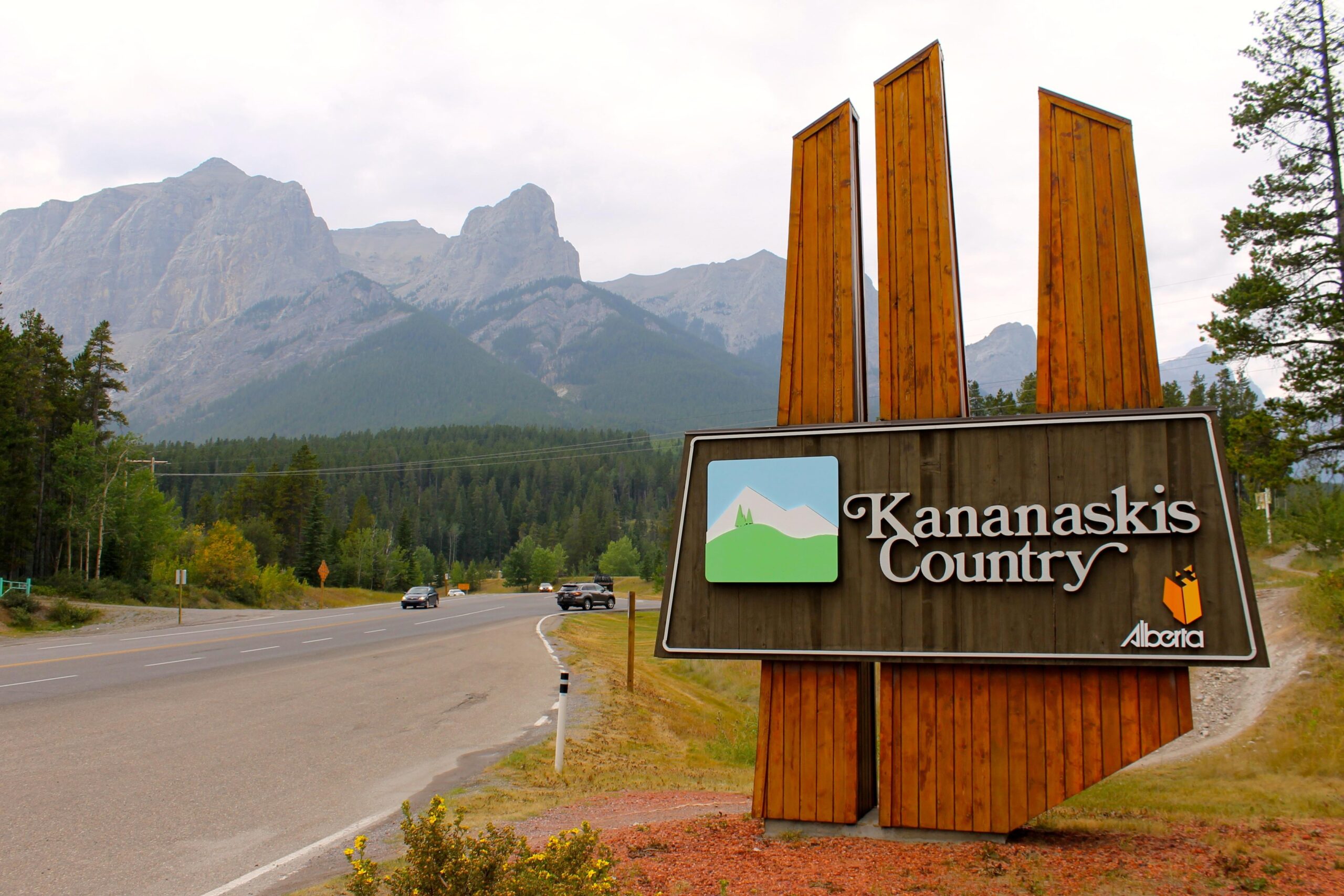 A sign for Kananaskis Country in Alberta stands by the roadside, featuring mountains in the background. The sign is designed with wooden elements and includes a graphic depicting mountains and a tree, along with the text "Kananaskis Country" and "Alberta." Lush green trees and a cloudy sky are visible around the area. Canmore Nordic Centre mountain bike trail.