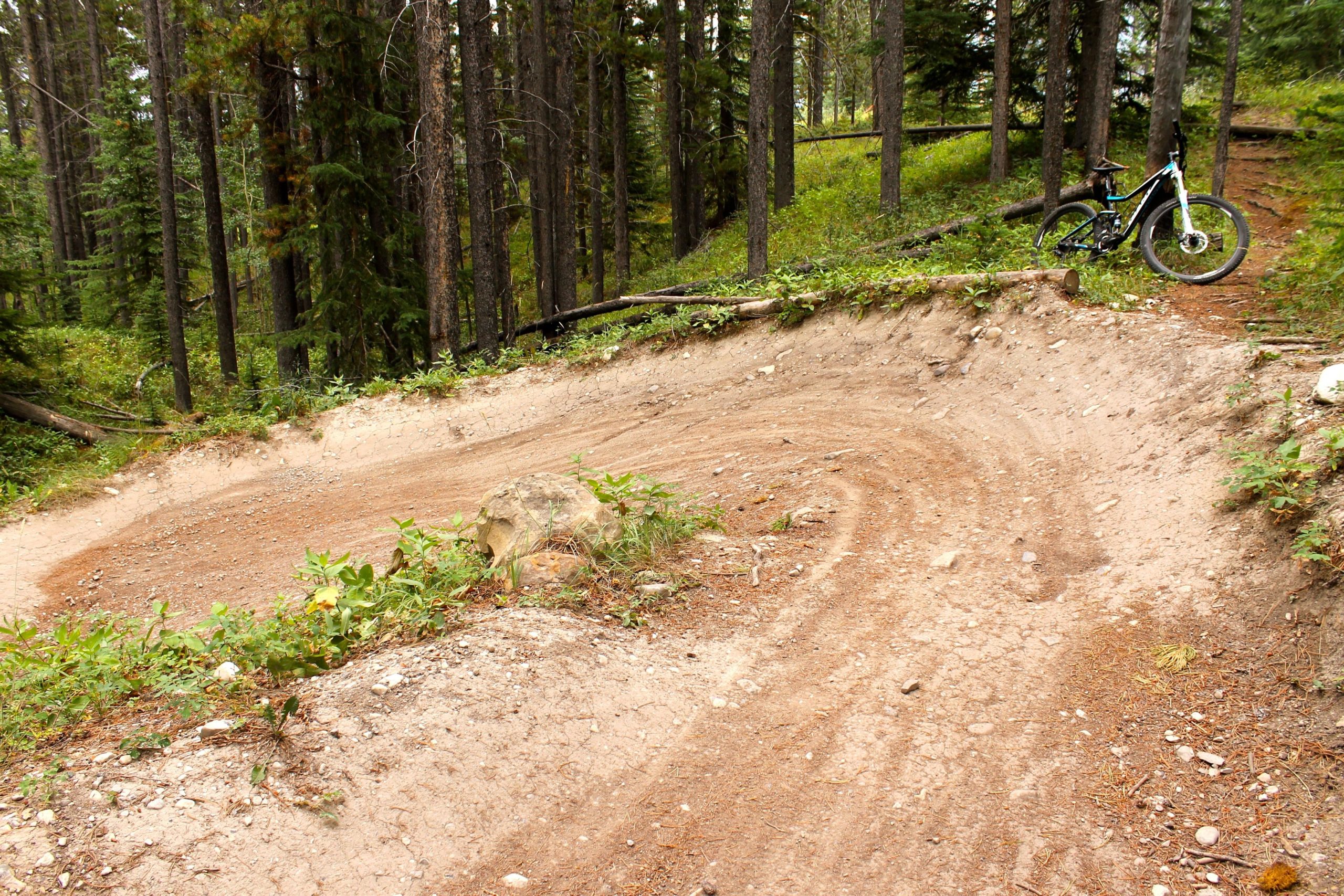 A winding dirt mountain bike trail through a forested area, featuring a curved path and a mountain bike resting on the side. Surrounding vegetation includes small bushes and pine trees, with a few rocky elements visible on the trail. Canmore Nordic Centre mountain bike trail.