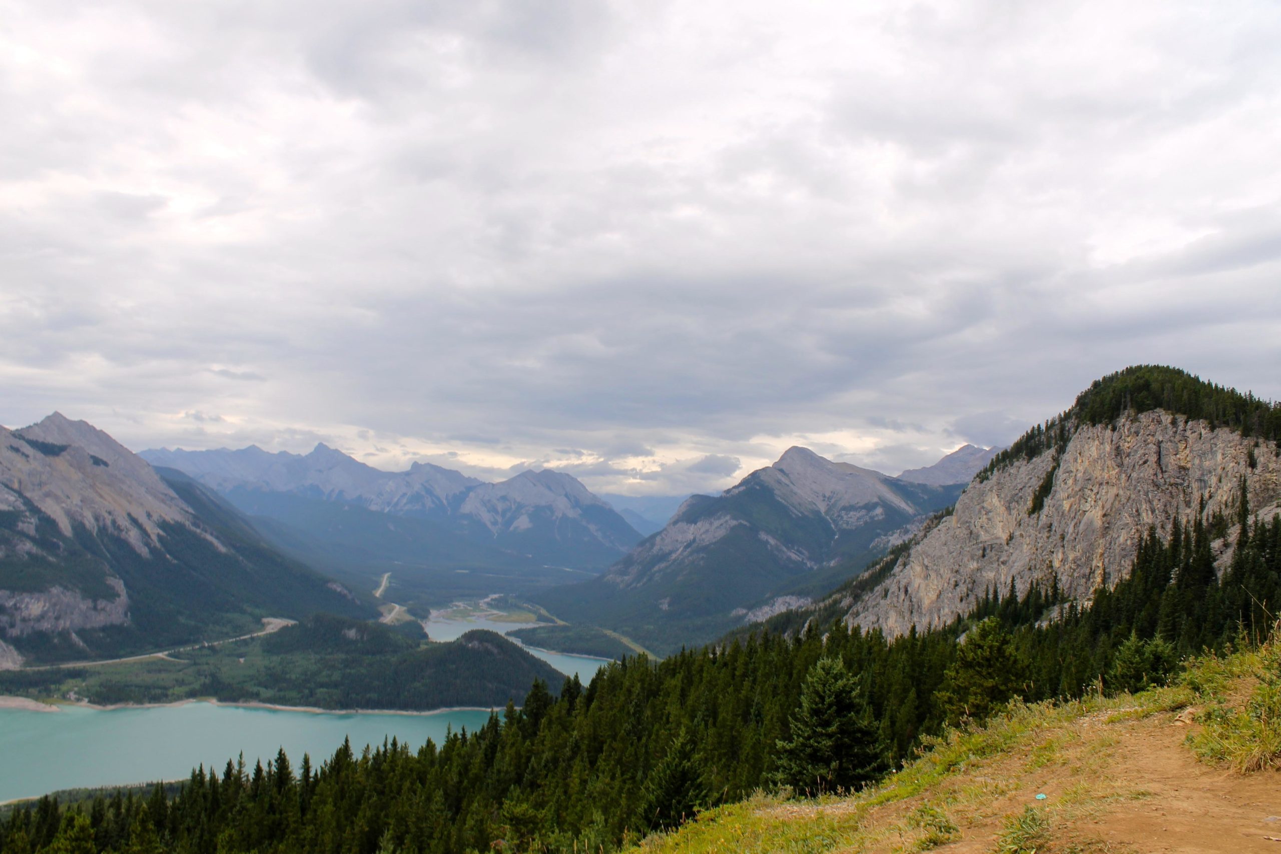 A panoramic view of a mountainous landscape under a cloudy sky, featuring rugged mountains, a winding river, and a forested foreground. The scene captures a mix of deep greens and earthy tones, with various peaks in the distance. Prairie View mountain bike trail.