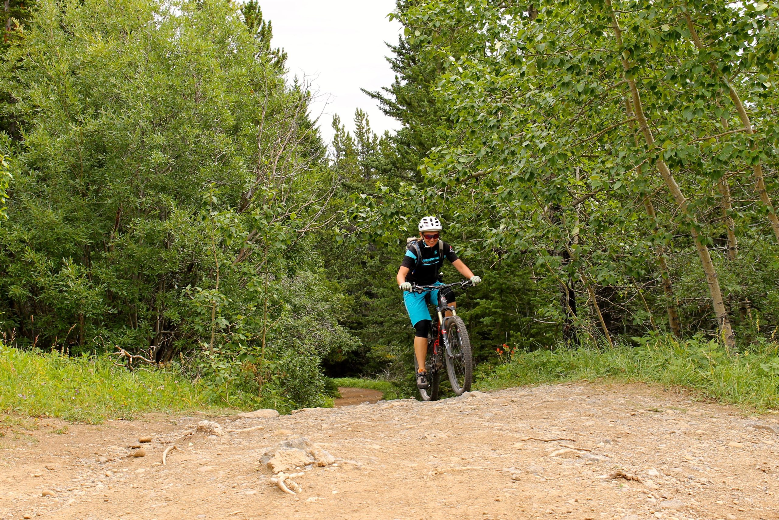 A person riding a mountain bike on a dirt trail surrounded by lush green trees. The cyclist is wearing a helmet and athletic gear, navigating over a rocky section of the path. Prairie View mountain bike trail.