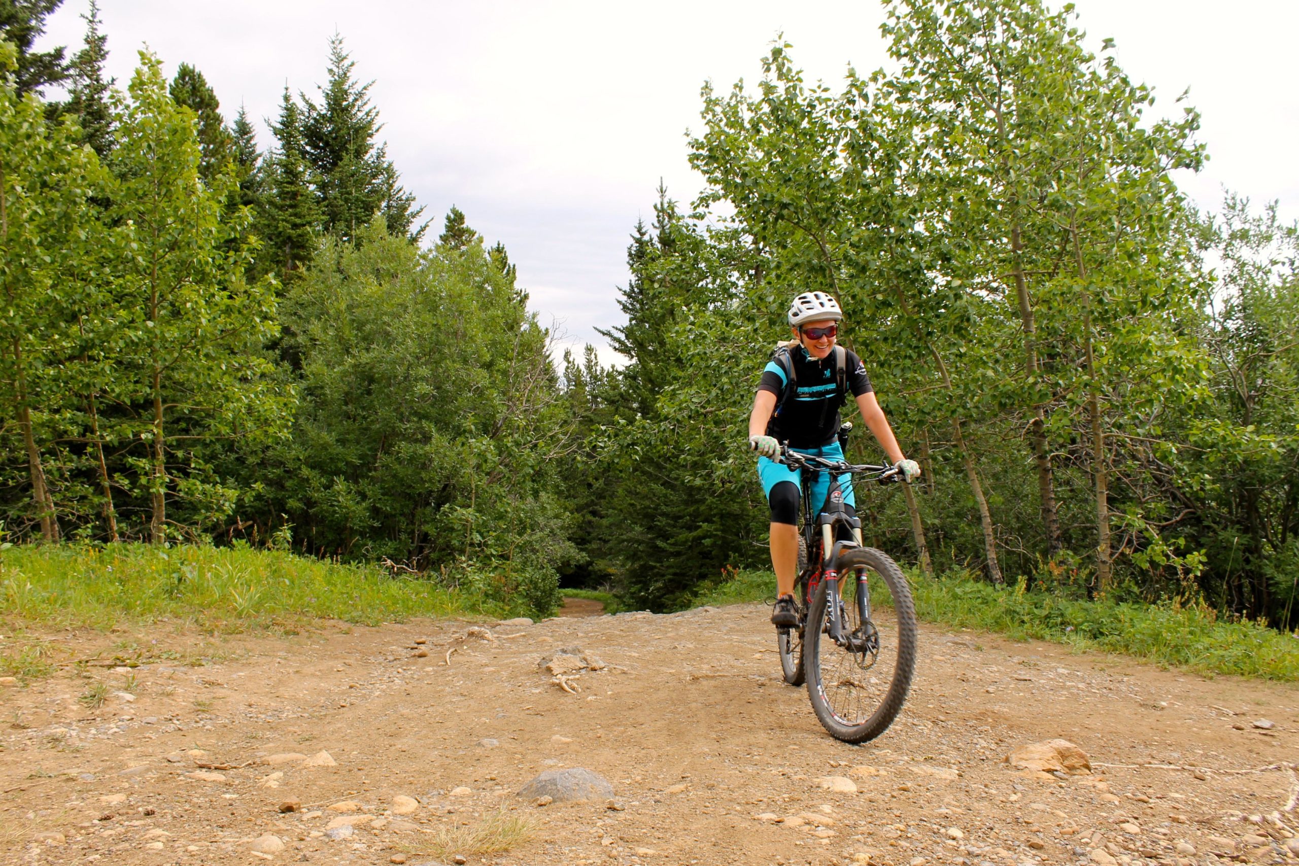 A mountain biker riding on a dirt trail surrounded by green trees and foliage. The rider is wearing a helmet and sports attire, navigating a natural, rugged path amidst a scenic outdoor setting. Prairie View mountain bike trail.