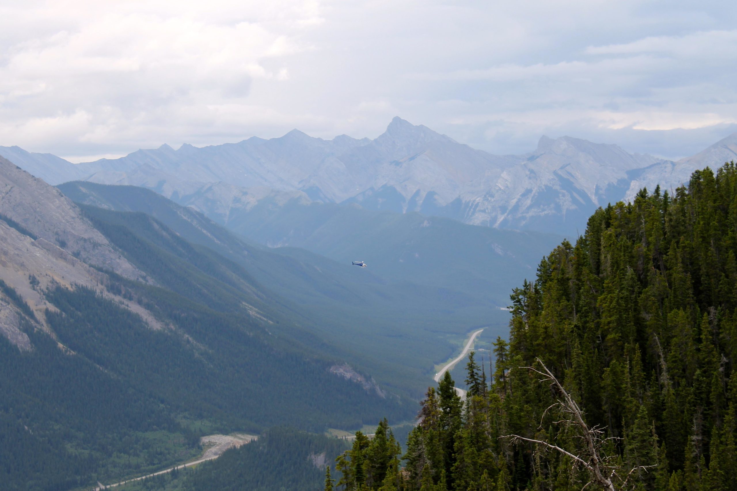 A panoramic view of a mountainous landscape featuring rugged peaks in the background, lush green valleys, and a winding road. A helicopter is visible flying in the distance against the cloudy sky. Foreground includes dense coniferous trees. Prairie View mountain bike trail.
