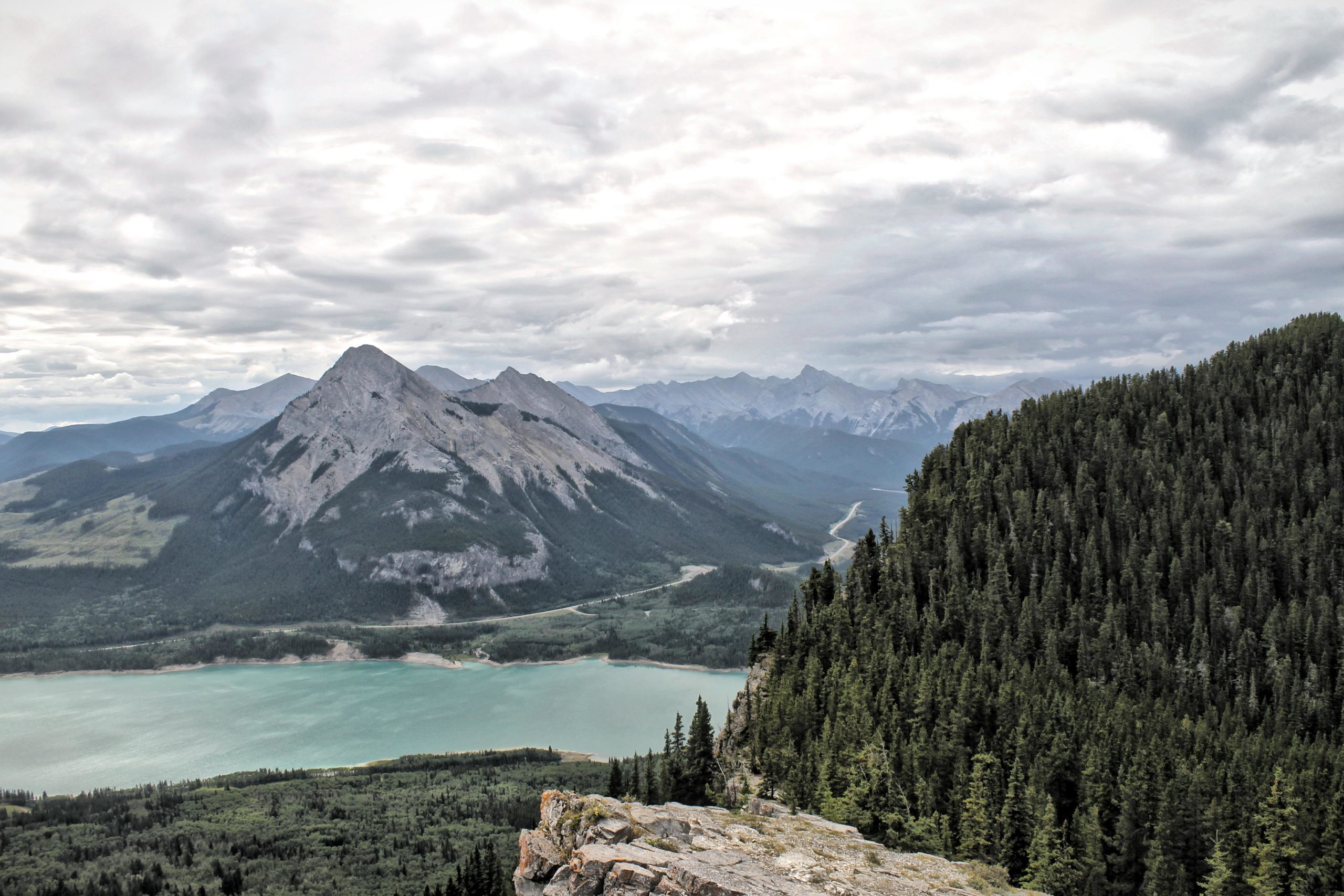 A panoramic view of mountain ranges under a cloudy sky, featuring steep rocky peaks on the left and dense evergreen forests on the right. Below, a calm turquoise lake is surrounded by greenery, while a winding road cuts through the valley. Prairie View mountain bike trail.
