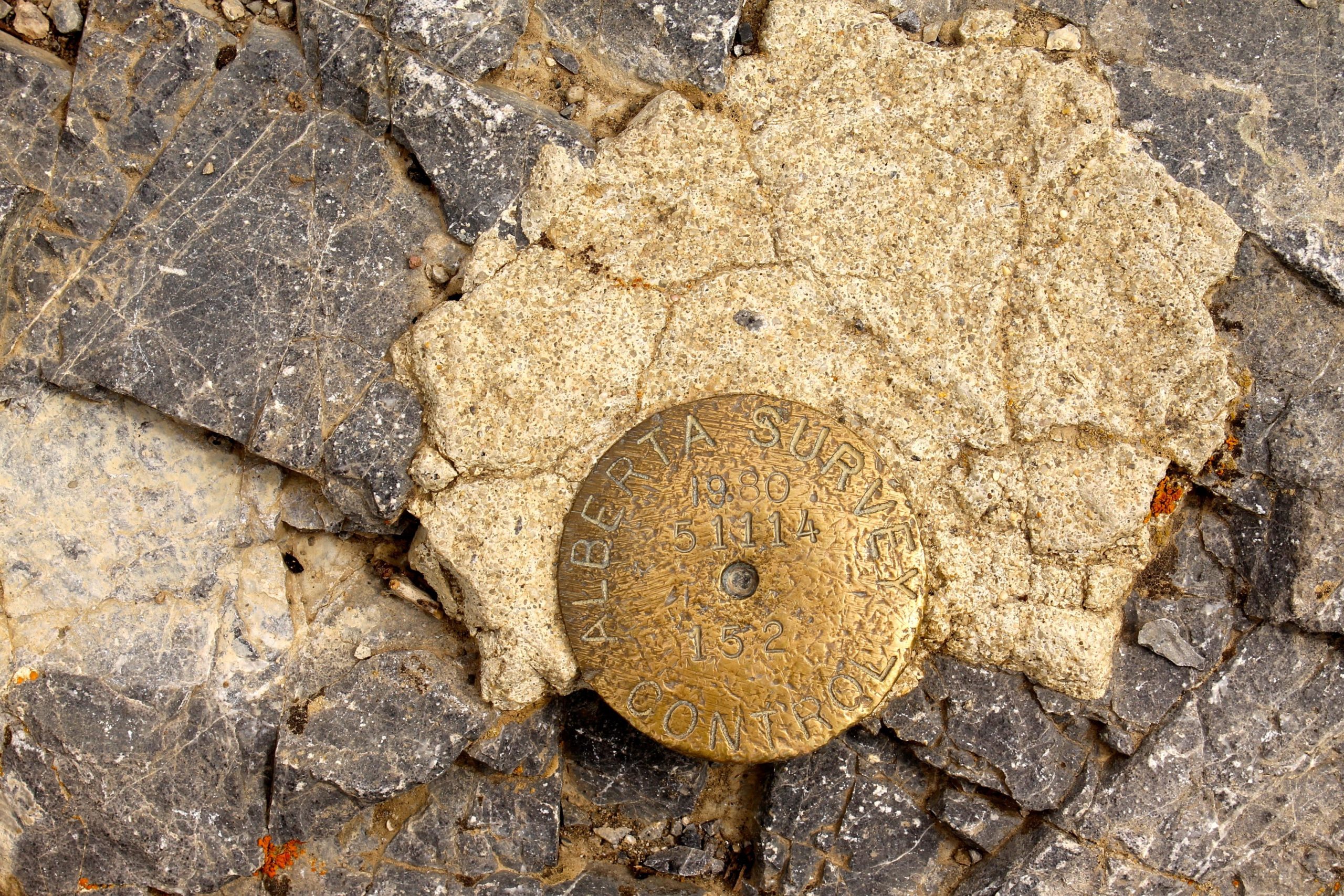 A metal survey marker embedded in rocky terrain, labeled "ALBERTA SURVEY 1980 51114 CONTROL 152," surrounded by various shades of gray and yellowish stones. Prairie View mountain bike trail.