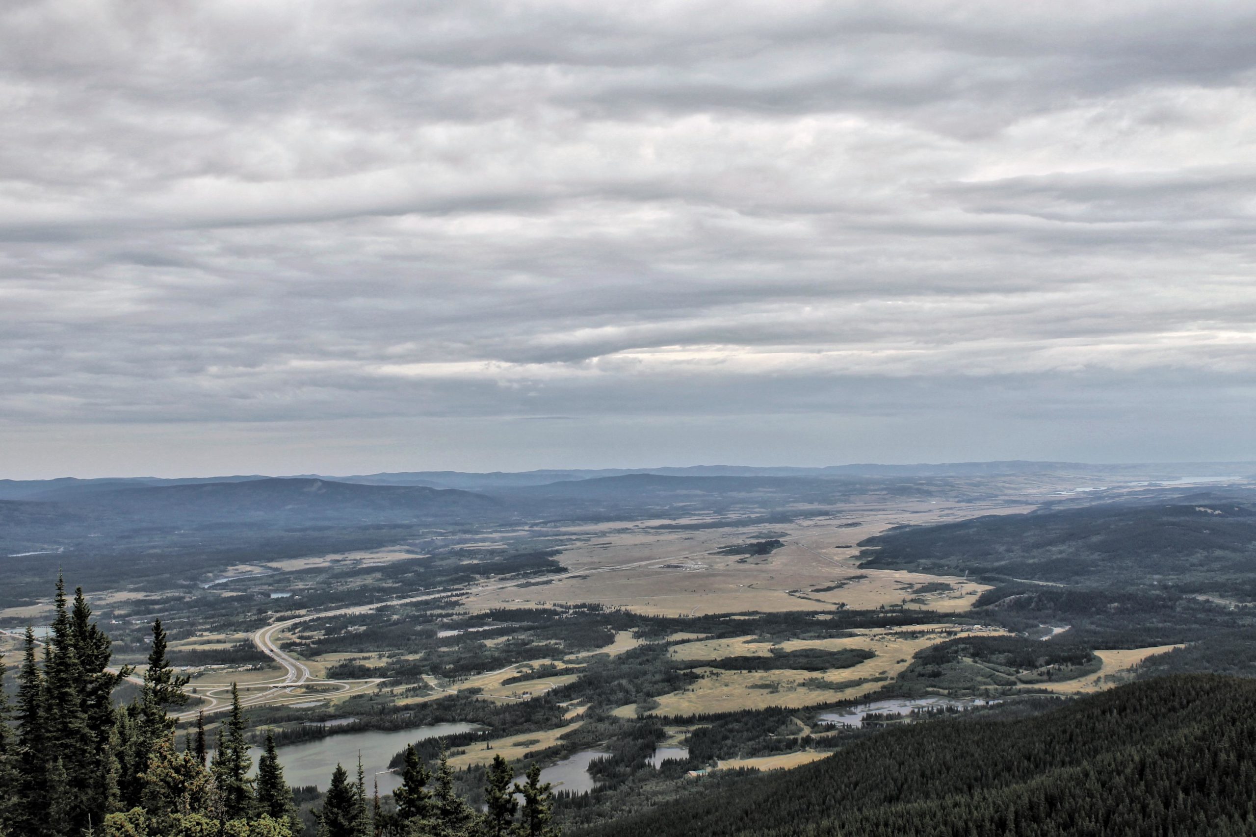A panoramic view of a valley surrounded by mountains under a cloudy sky, featuring winding rivers and patches of greenery interspersed with open fields. Prairie View mountain bike trail.