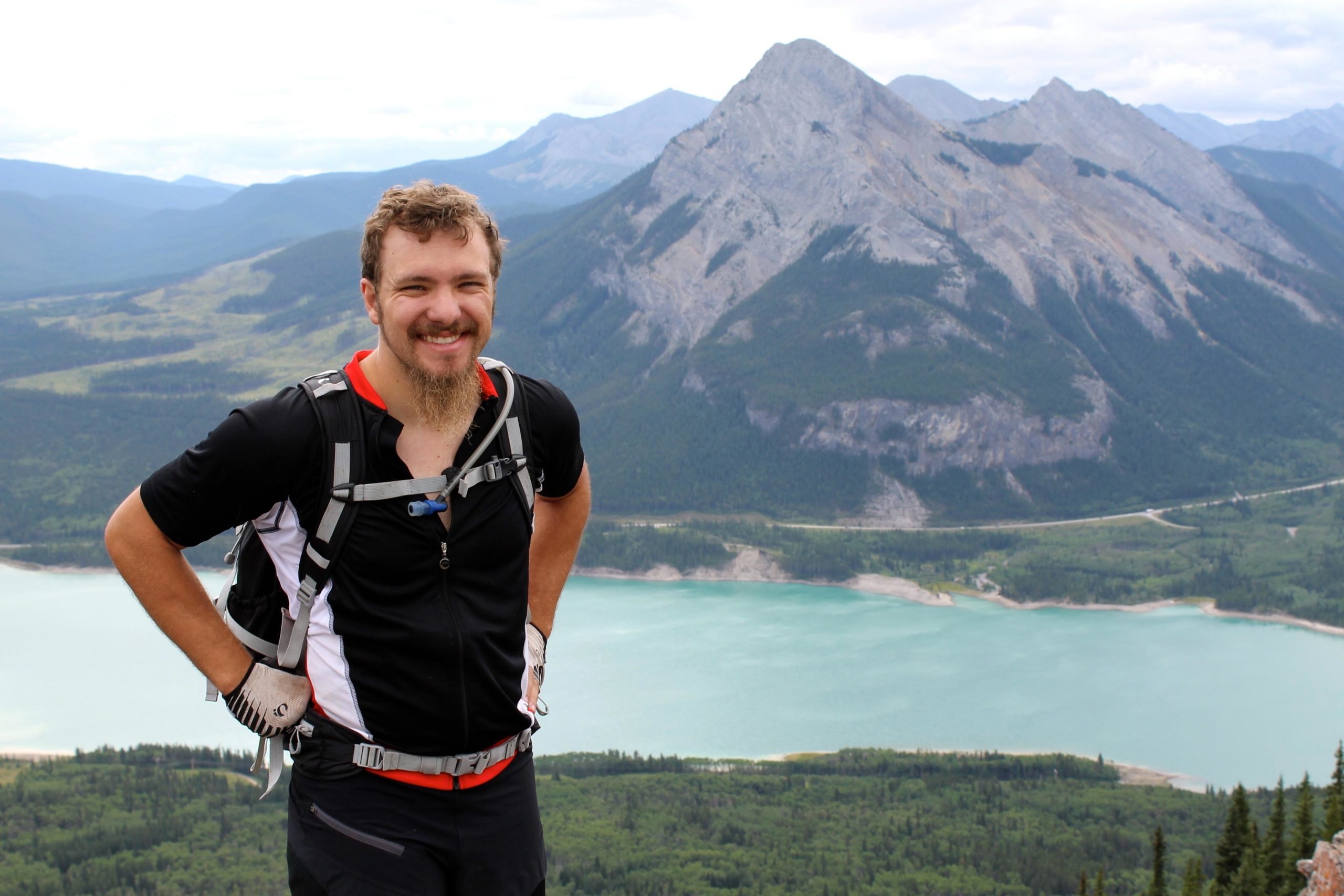 A person smiling while standing atop a mountain, wearing a black and red athletic shirt and a backpack. The background features a scenic view of green valleys, a turquoise body of water, and rocky mountain peaks under a partly cloudy sky. Prairie View mountain bike trail.