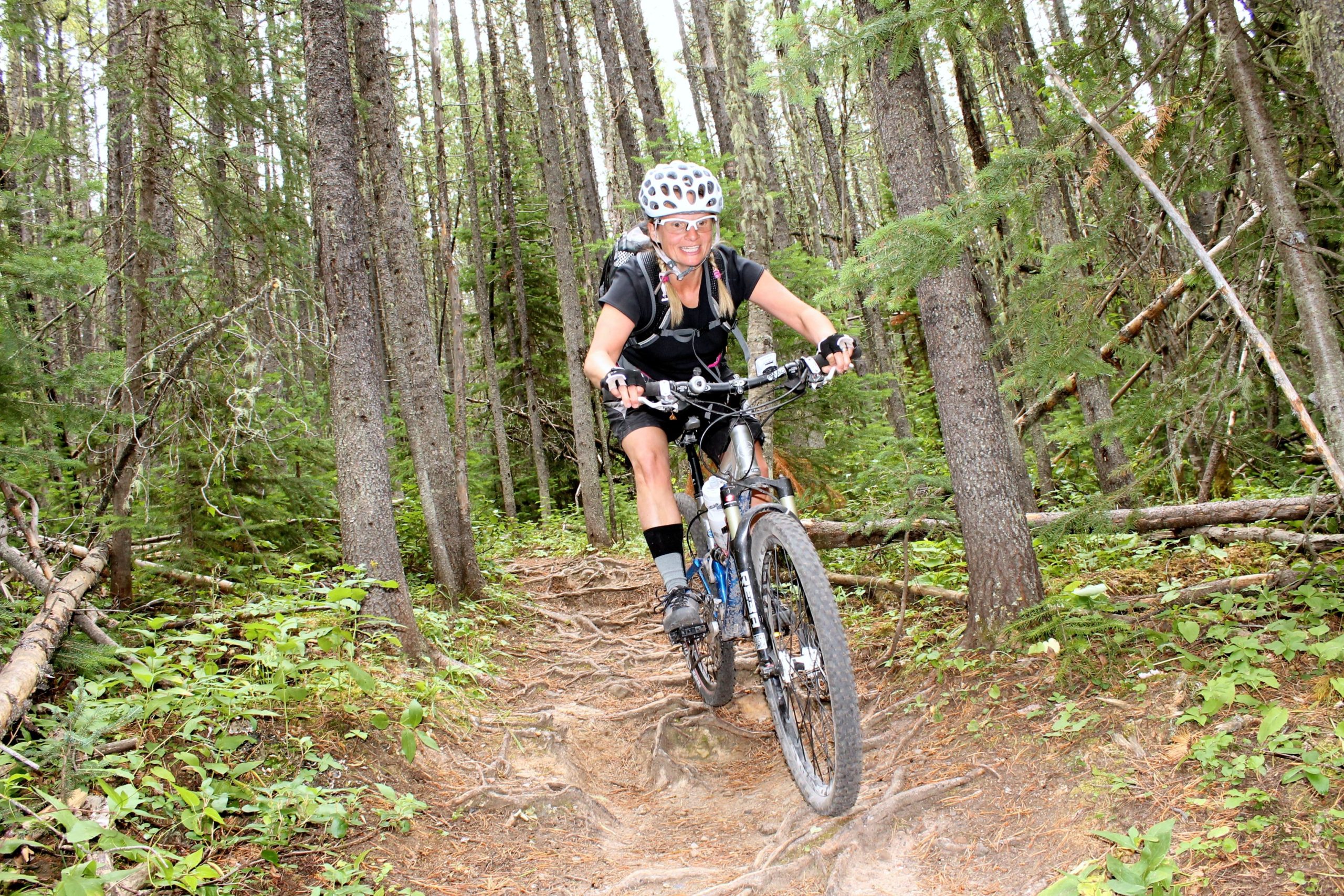A person riding a mountain bike on a narrow, rustic trail surrounded by tall trees and greenery. The cyclist is wearing a helmet and is actively navigating through a challenging section of the path, which features exposed tree roots and uneven terrain. Prairie View mountain bike trail.