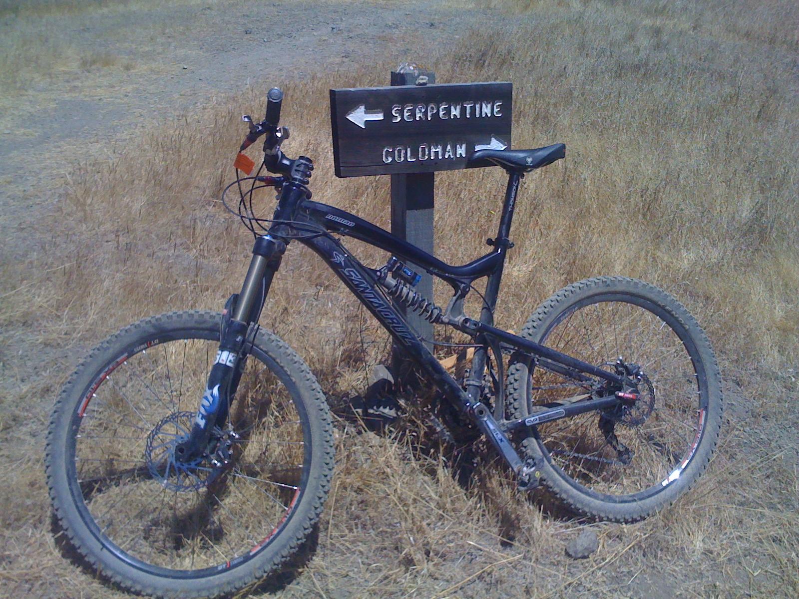 Santa Cruz Nomad: Mountain bike leaning against a wooden signpost indicating trail directions for "Serpentine" and "Goldman," set in a dry, grassy landscape.