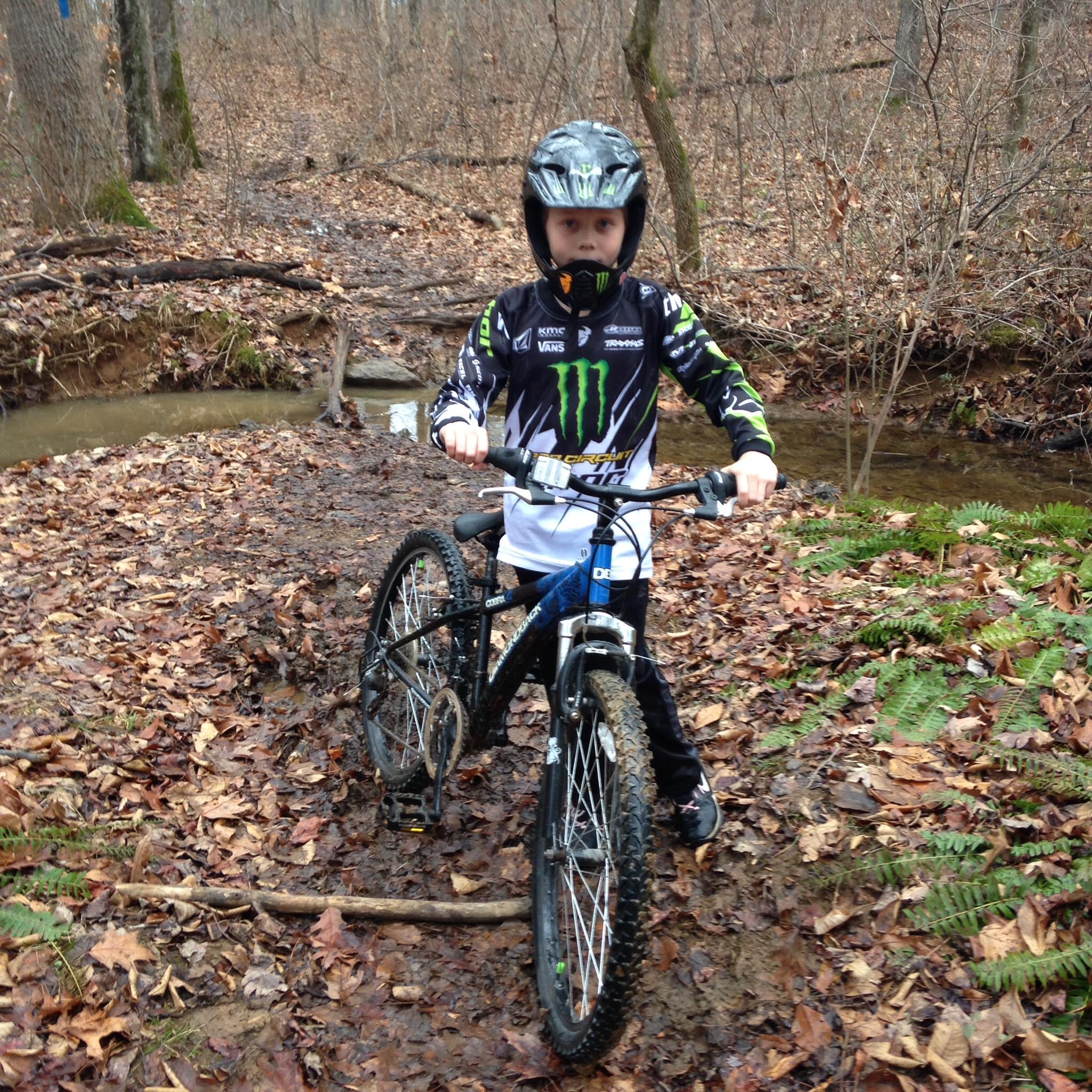 A young child in a black and white motocross jersey and helmet is standing next to a mountain bike in a wooded area covered with fallen leaves. Behind them, a shallow stream can be seen, and the surrounding landscape features bare trees and ferns. The child looks focused and ready for an adventure. Whitney State Forest mountain bike trail.