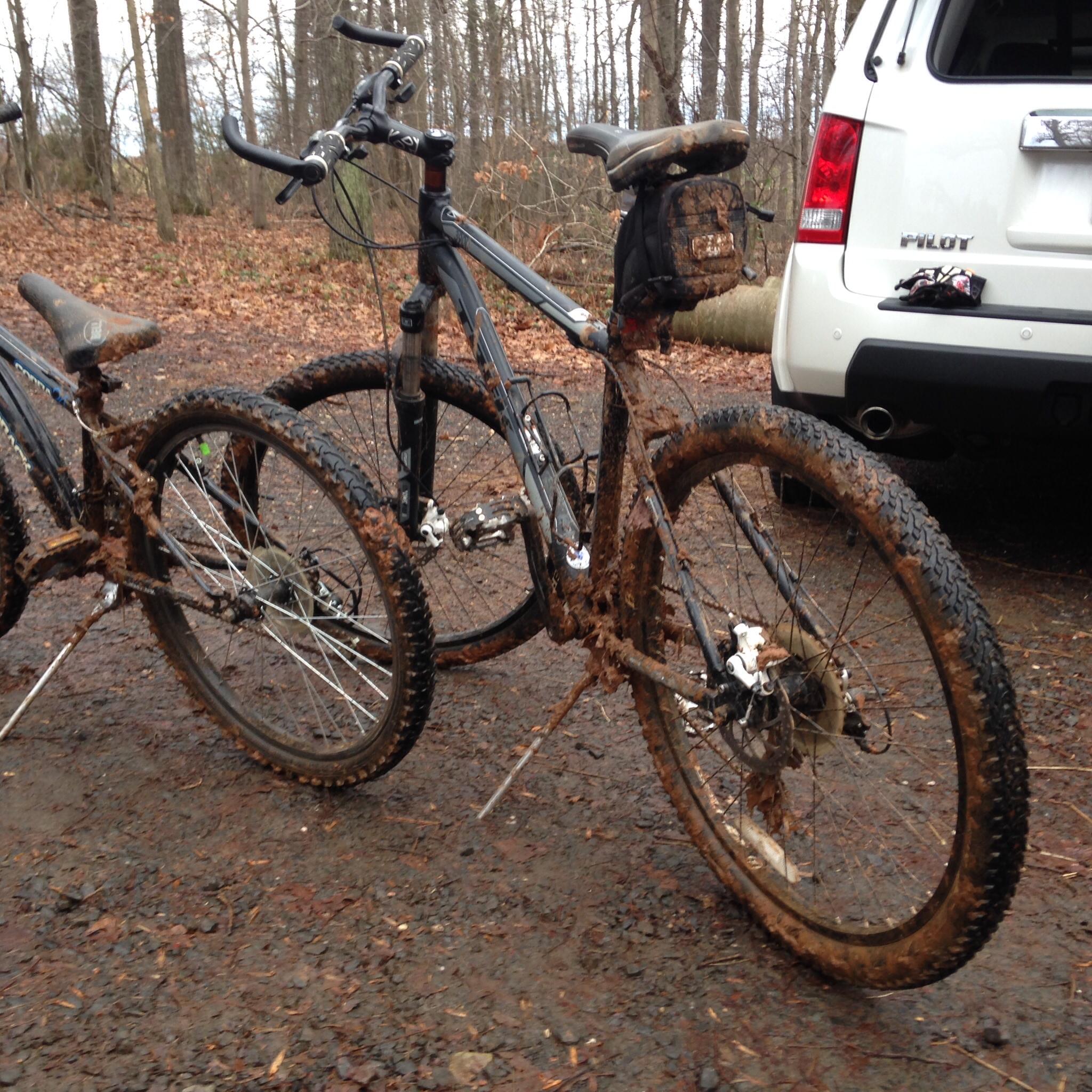 Two mountain bikes are parked on a muddy trail, covered in mud from recent use. The bikes are positioned closely together, showcasing their dirty tires and frames. In the background, there is a white SUV parked, surrounded by leafless trees, indicating a forested area in an outdoor setting. Whitney State Forest mountain bike trail.