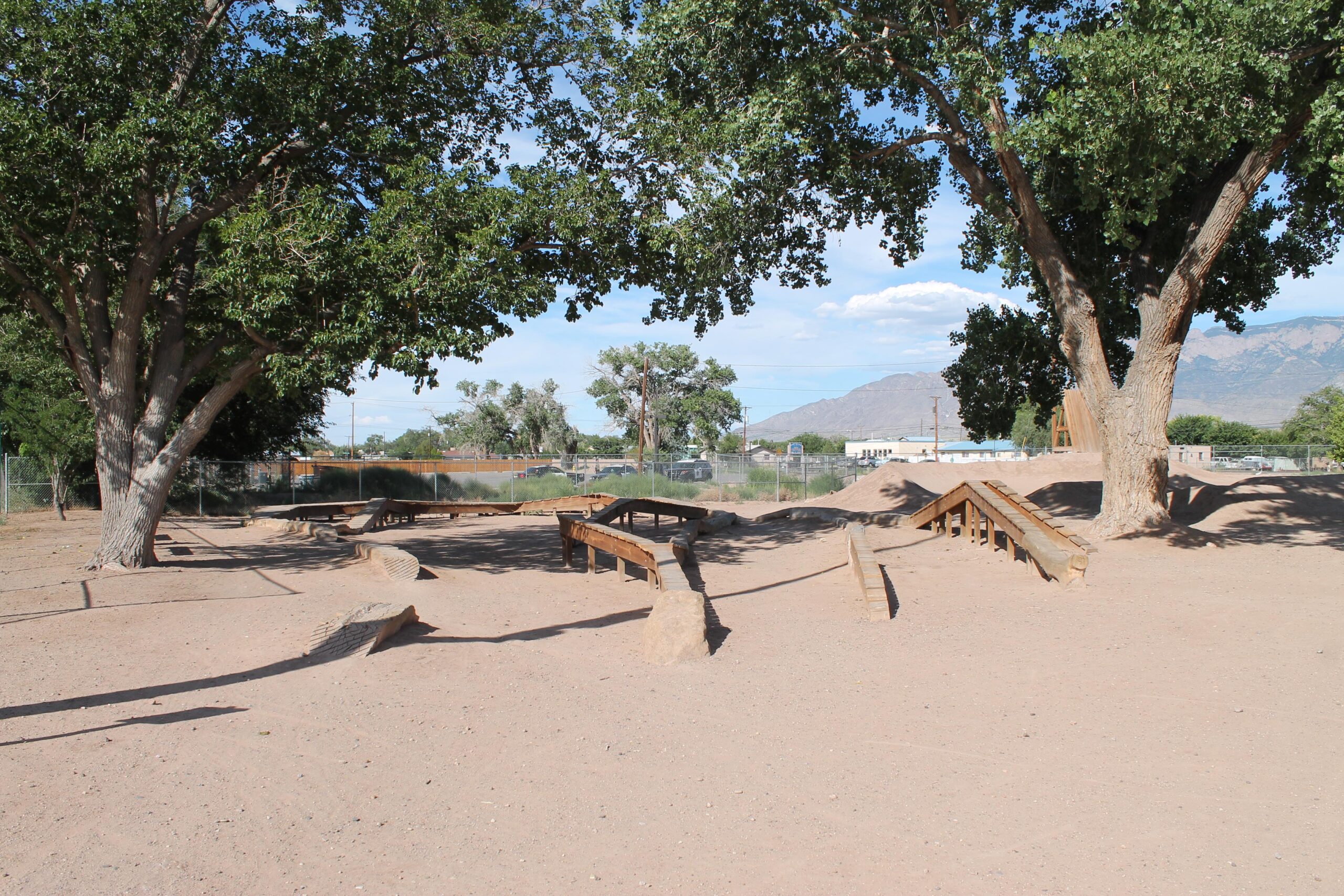 A sandy play area surrounded by large trees, featuring wooden structures and ramps for outdoor activity. In the background, distant mountains are visible under a partly cloudy sky. North Valley Bike Park mountain bike trail.