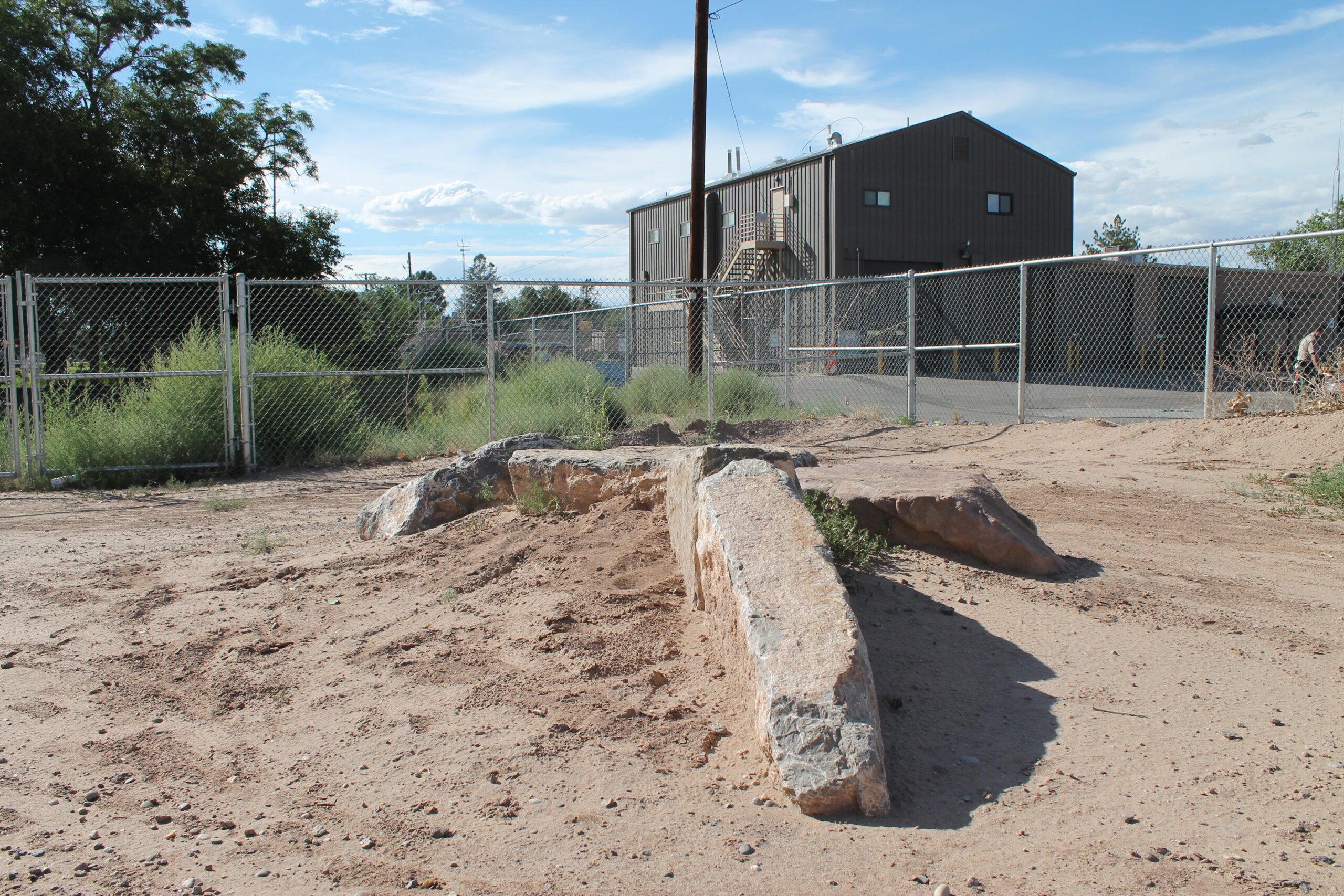A sandy area with a large, weathered rock formation partially covered in dirt and small plants, in front of a fenced facility. A building with a metal exterior is visible in the background, along with a power pole and a clear blue sky scattered with clouds. North Valley Bike Park mountain bike trail.