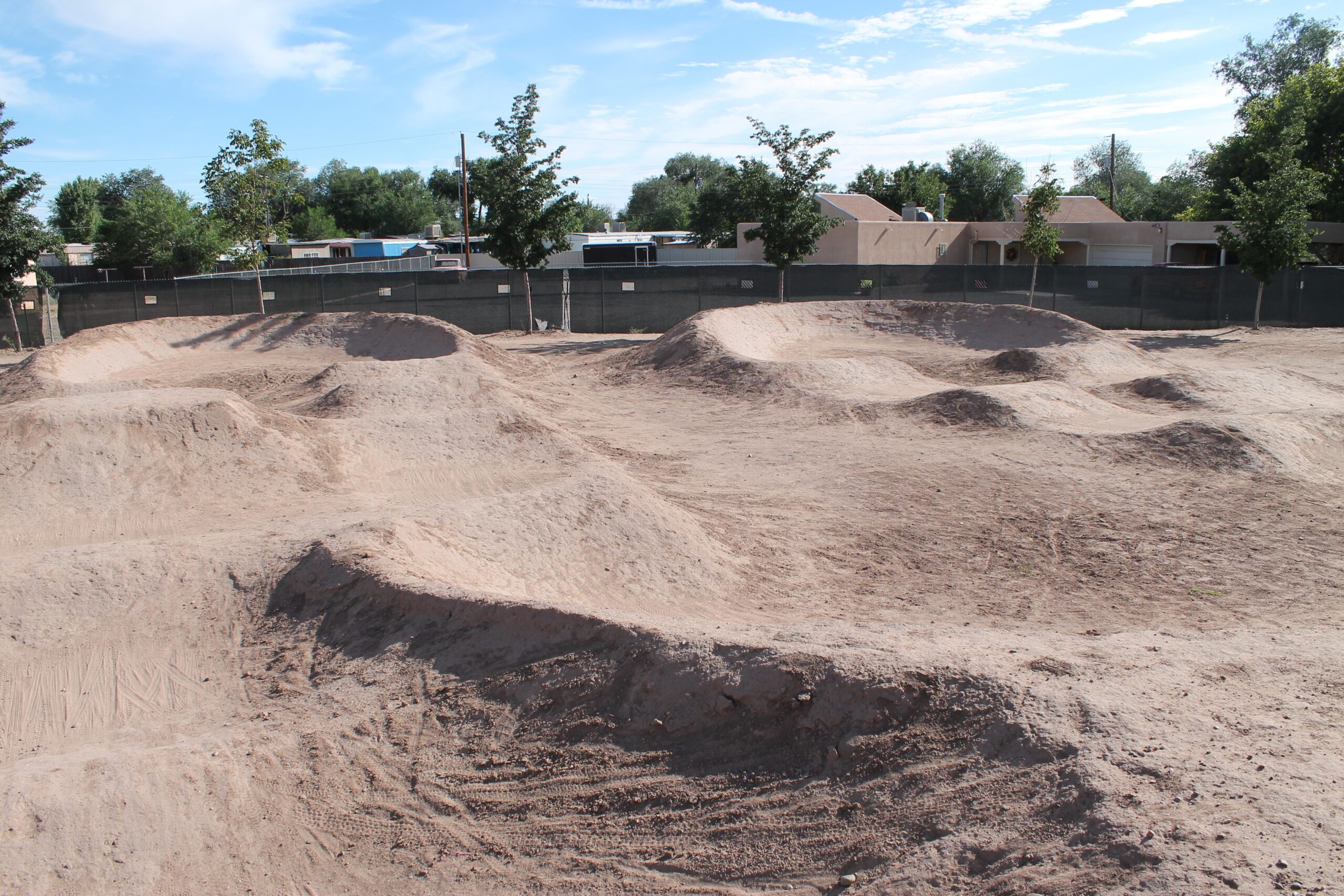 A dirt bike track featuring several mounds and jumps, surrounded by trees and a fence, with a clear blue sky in the background. North Valley Bike Park mountain bike trail.
